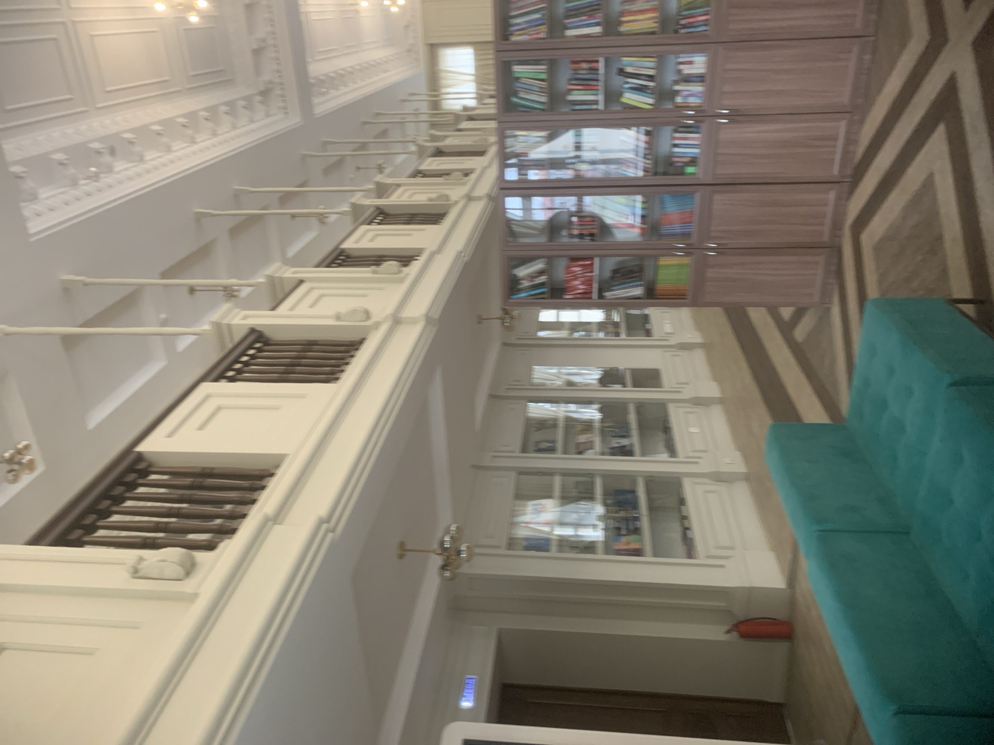 This indoor scene, likely in Izhevsk, Russia, shows a library interior featuring high ceilings with ornate molding, white walls, and wooden bookshelves stocked with books. Some sections have glass-fronted doors. A teal sofa sits alongside the shelves. The floor has a patterned design. An extinguisher is visible on the wall.