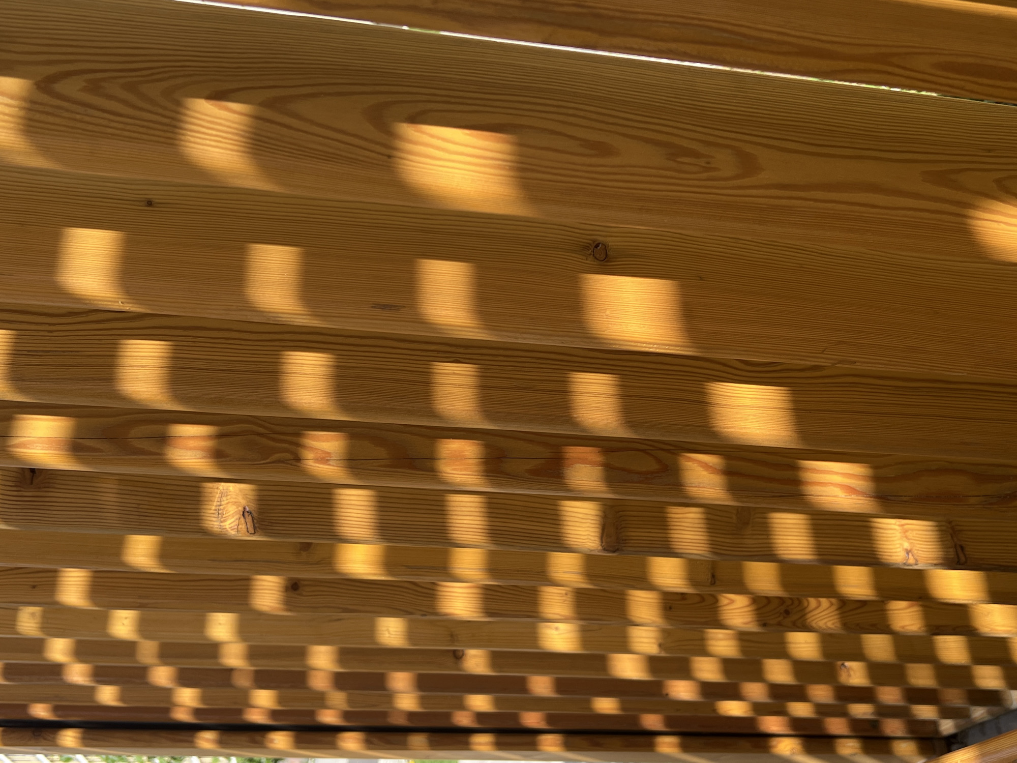 The image shows a close-up of the underside of a wooden structure, likely a deck or a pergola, with sunlight creating a pattern of alternating light and shadow across the wooden planks. The wood appears to be light-colored, possibly pine or a similar softwood, and the grain is visible. The lighting suggests a sunny day. The location in Yekaterinburg, Russia is not discernible from the image. There are no people present in the image.