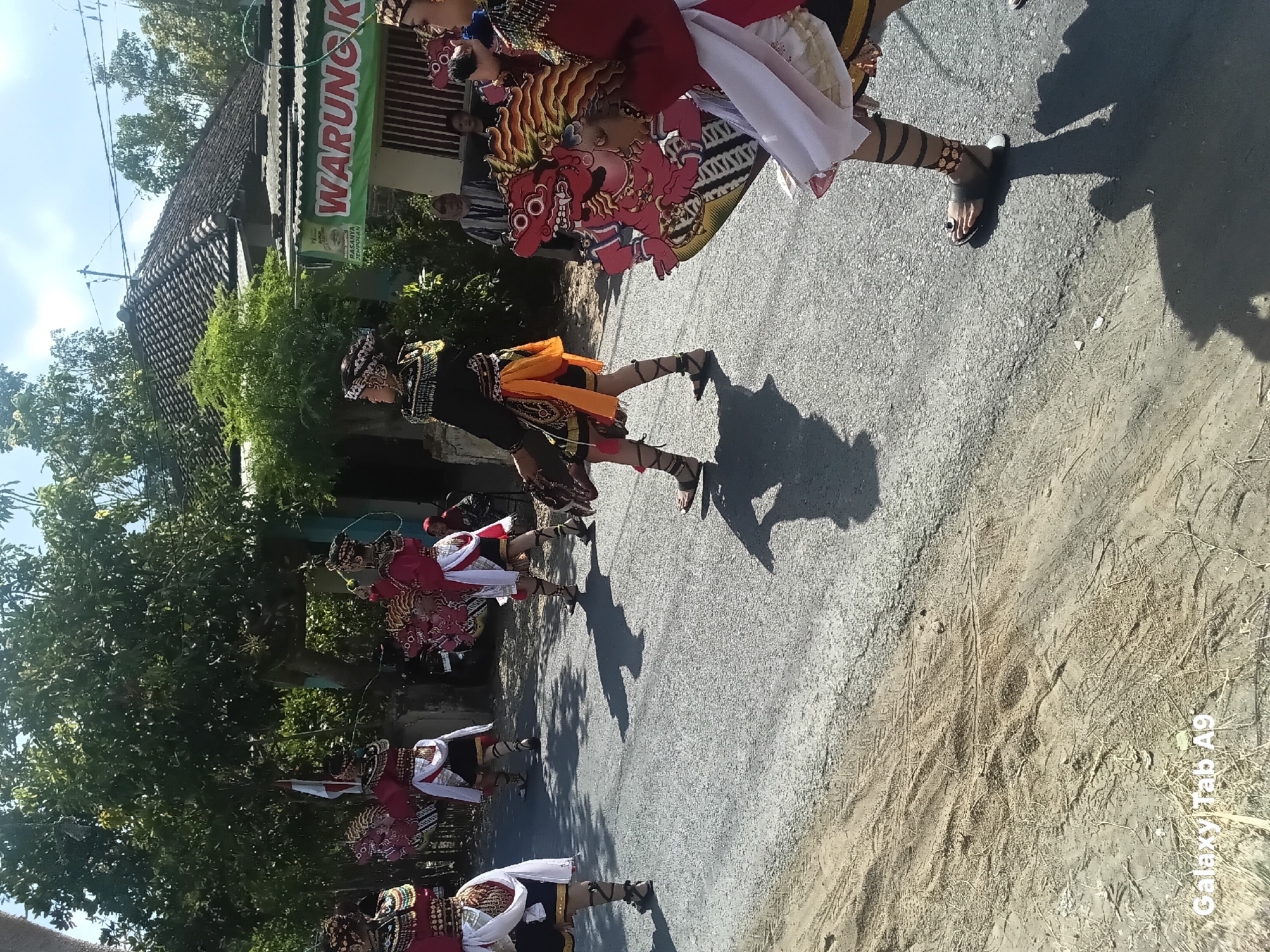 In Bendorejo, Indonesia, a procession is taking place on a paved road. Several individuals are dressed in elaborate, traditional costumes featuring vibrant red and black fabrics, gold accents, and decorative headwear. Some participants are carrying large, colorful effigies that appear to be mythological figures with red faces and elaborate headdresses. The figures are adorned with intricate patterns. The procession is moving along the road, casting shadows on the asphalt. In the background, there are houses with tiled roofs and lush green trees. A sign for "WARUNG KOP" is visible, indicating a local establishment. The environment appears to be sunny and warm, with a clear blue sky.