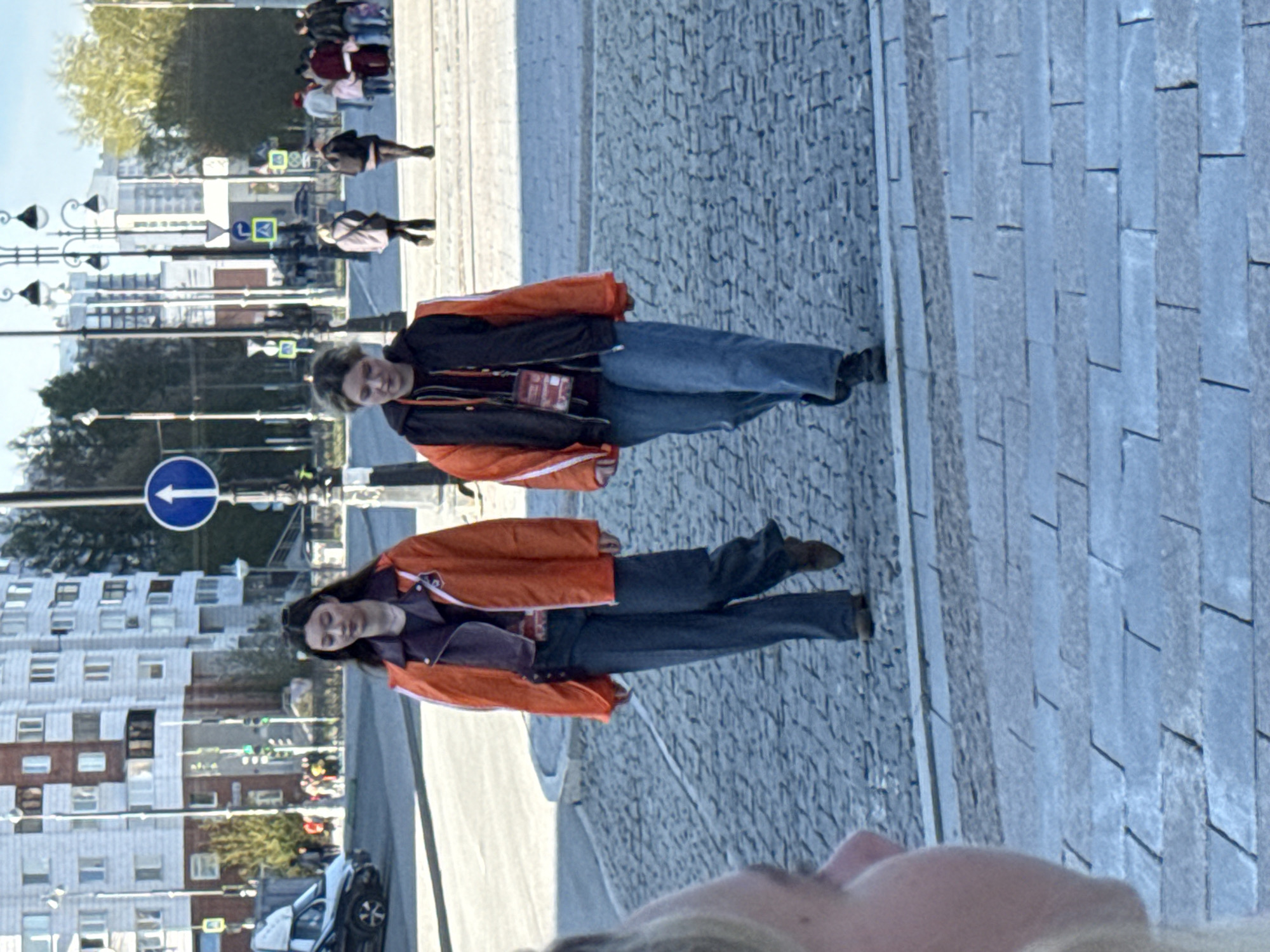 The image shows two people standing on a cobblestone street, likely a pedestrian area, in what appears to be a city. They are both wearing orange and black jackets and jeans. One person is slightly ahead of the other. Buildings are visible in the background, as well as street signs and other people further away. The street is lined with streetlights, and there are parked cars and traffic signs present. Based on the urban architecture and the style of street signs, the location is likely Yekaterinburg, Russia.