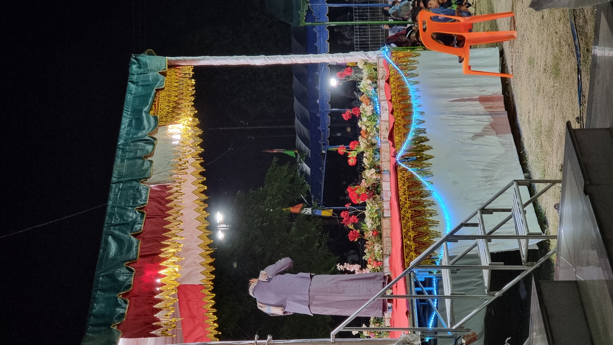 The image depicts an outdoor evening event at Tumpokbeurandang, Indonesia, likely a cultural performance or ceremony. A man, dressed in a traditional light purple long coat and sarong, stands on a decorated stage, facing away from the camera, with his hands raised as if in prayer or a gesture of greeting. The stage is adorned with red fabric, ornate gold trim, and illuminated by blue string lights along its front. Red and white flowers are arranged as a border on the stage. Behind the stage, a blue tent structure is visible, with a crowd of people gathered, some sitting and watching. To the right of the stage, an empty orange plastic chair is placed on the ground. The background consists of trees and the night sky. The overall atmosphere suggests a community gathering or celebration.