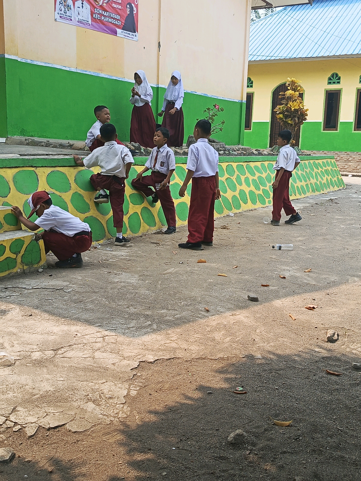 In an outdoor schoolyard in Karya Dadi, Indonesia, several young students in uniforms are engaged in various activities. The school building features a beige upper story and a green lower story, with a yellow wall adorned with green circular patterns. Several children, dressed in white shirts and maroon pants or skirts, are scattered across the scene.
In the foreground, a boy wearing a red cap and white shirt is crouched down, appearing to clean or examine the base of a yellow and green patterned wall. To his right, another boy in a white shirt and maroon pants is seated on a low step. Further back, near the wall, a boy in a white shirt and maroon pants stands looking away from the camera.
To the left, two girls wearing white hijabs and maroon skirts stand near the school building. One appears to be gesturing with her hands, while the other looks down. In the middle ground, a boy in a white shirt and maroon pants is perched on the patterned wall, looking towards the right. Another boy, also in a white shirt and maroon pants, is walking away from the camera towards the right. On the ground in front of him, a plastic bottle and a clear plastic cup are visible.
The ground is a mixture of concrete and gravel, with scattered leaves and small stones. The overall environment suggests a typical school setting during daylight hours.