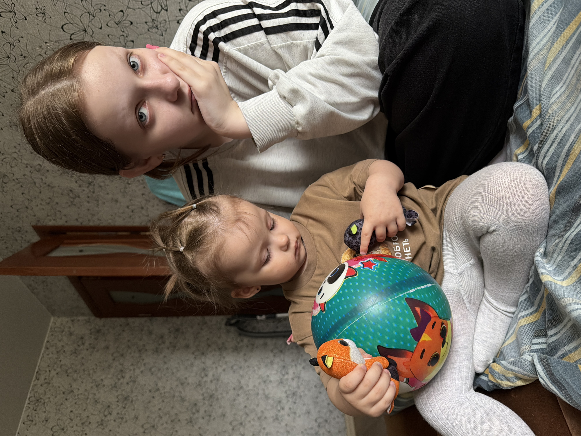 Two people are captured in an indoor shot: a teenager and a toddler. The teenager is positioned at the top of the frame, wearing a white hooded sweatshirt with black stripes on the sleeves and black pants. Her hand is partially covering her face, and she is looking directly at the camera. Below her, a toddler is lying down. The toddler is dressed in a tan shirt and white tights and is clutching a colorful ball with cartoon animal designs and a small plush toy. The background reveals a wallpapered wall and a striped blanket or throw, suggesting the location is a residential setting. The composition implies that the photo was taken in Razdolie, Russia.