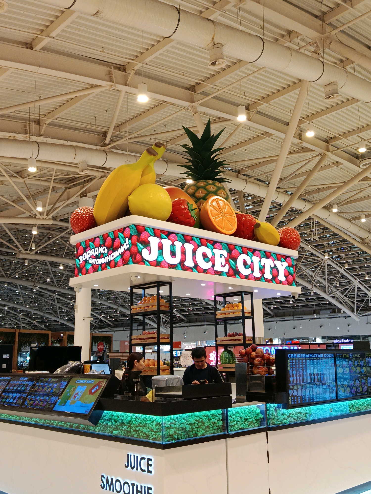 A brightly lit juice bar kiosk, named "JUICE CITY," is prominently displayed with an overhead canopy adorned with oversized artificial fruits, including bananas, a lemon, strawberries, an orange slice, and a pineapple. The kiosk's facade features a digital screen displaying fruit patterns and the text "JUICE CITY" in large white letters. Below this, a counter is equipped with multiple digital menus showcasing various juice options and prices, with two employees visible behind the counter. One employee, a woman, appears to be interacting with a customer, while the other, a man, is looking at his phone. The environment suggests an indoor setting, possibly a shopping mall or airport terminal, indicated by the high ceiling with exposed industrial-style ductwork and lighting fixtures. The kiosk is surrounded by other businesses, and a digital sign for "Play Today" is visible in the background. The overall scene depicts a commercial space focused on the sale of fresh juices and smoothies.