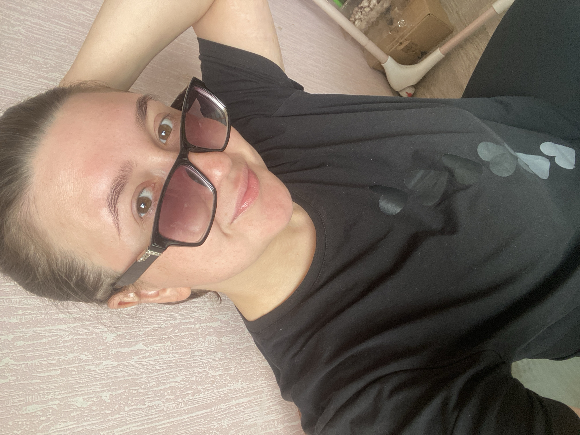 A young woman, positioned in the foreground, is captured in a selfie. She has fair skin, brown eyes, and her dark hair is pulled back. She is wearing black-rimmed sunglasses with pinkish lenses and a black t-shirt with a pattern of dark hearts. Her right arm is raised, with her hand resting on the back of her head, and she is looking directly at the camera with a slight smile. The background consists of a textured, pale pink wall to her left and a light-colored floor with a pink and white cleaning tool and a cardboard box to her right. The overall setting appears to be indoors, possibly a residential area in Chaikovsky, Russia, given the context.