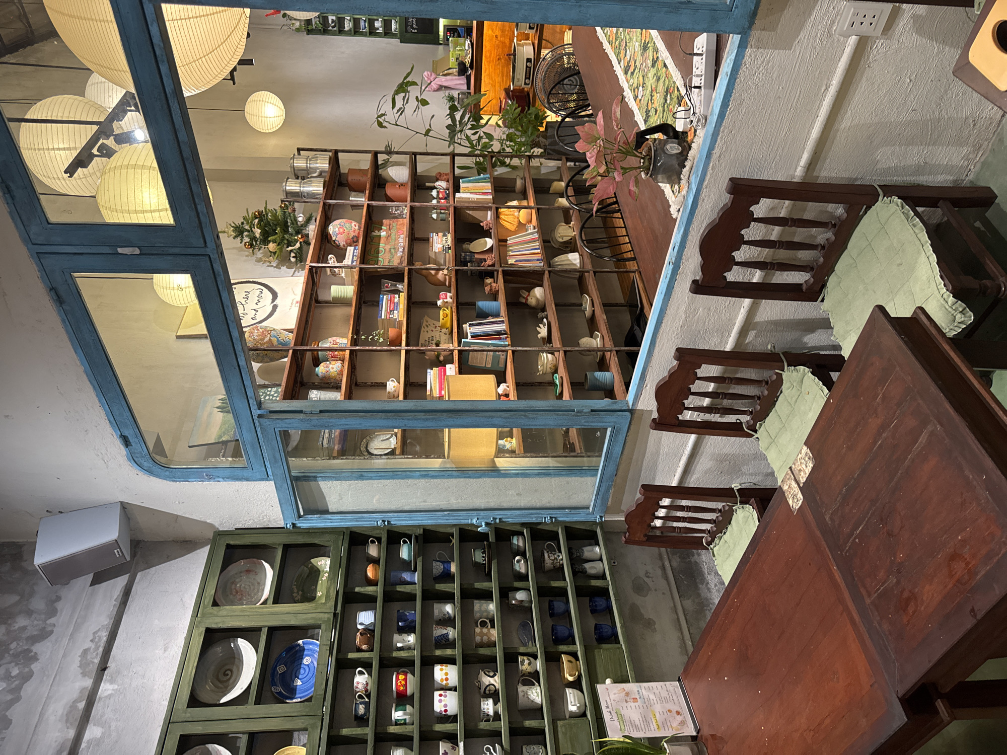 Indoor cafe in Đà Nẵng, Vietnam. Three dark wood chairs with light green cushions at a dark wood table with menus; built-in green display case with plates and mugs; a blue window frame looking at lanterns outside, bookshelf, and table.