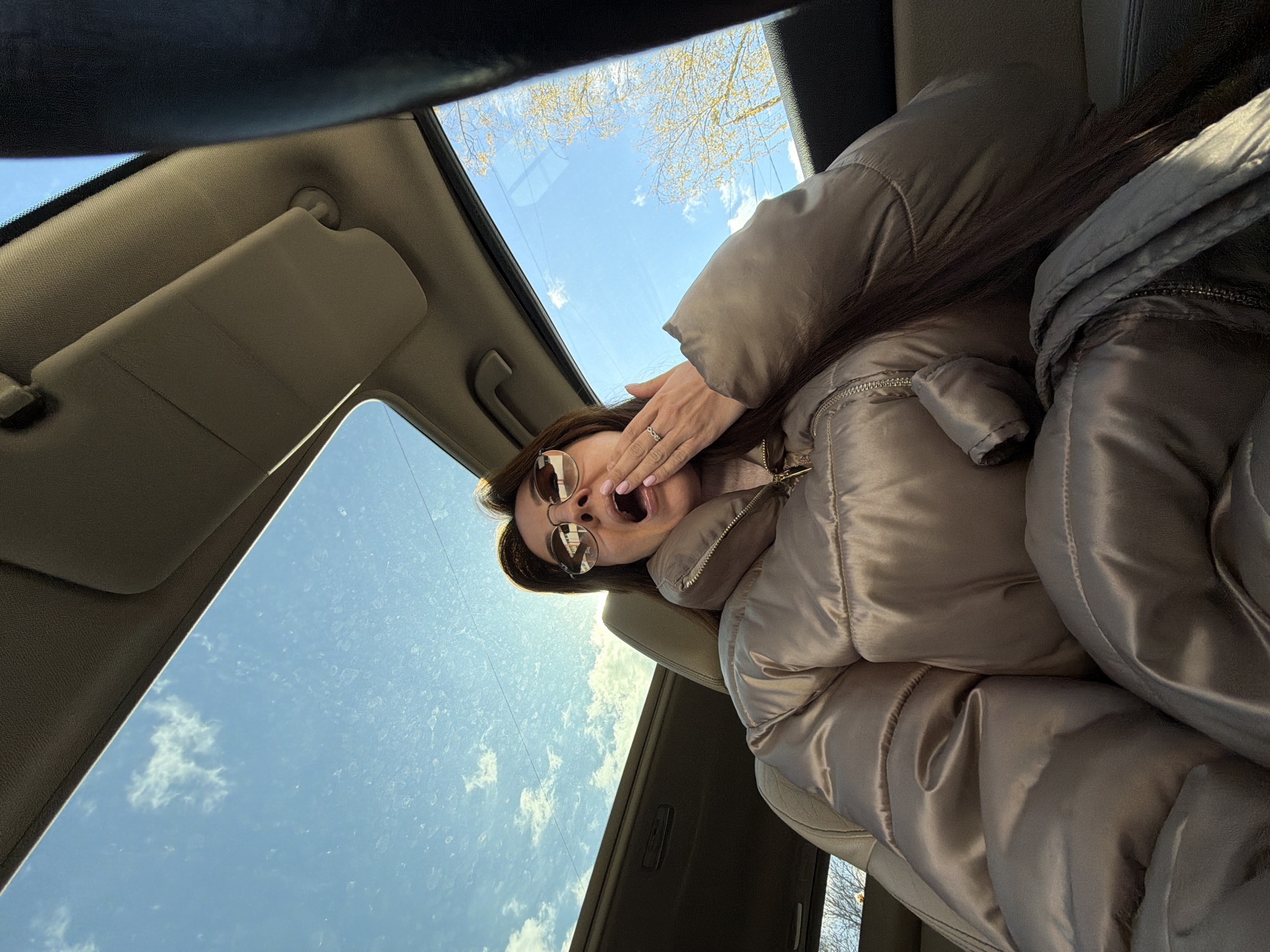 The image presents a unique, low-angle perspective from inside a car, looking upwards towards a woman and the sky through a sunroof. The woman is positioned diagonally, appearing to be in a passenger seat. She is the central subject, captured in a candid moment with her mouth wide open, suggesting a yawn or an exclamation. Her right hand is raised, with her fingers partially covering her cheek or head. A silver-toned ring is visible on her ring finger, and her fingernails are painted light pink.
She has long, dark hair and is wearing a voluminous, shiny, light brown or beige puffy winter jacket with a visible zipper and collar detail. Round, reflective sunglasses are perched on her face, obscuring her eyes.
The car's interior is visible around her, including the light-colored ceiling, a sun visor, and what appears to be the car's seat. Above and around her, the car's sunroof provides a view of the outside. Through the sunroof, a bright blue sky with scattered white clouds is prominent. Some smudges or speckles are visible on the sunroof glass. Bare tree branches are visible against the sky in the upper left portion of the image, suggesting a time of year like late autumn, winter, or early spring. The strong light indicates it is daytime and sunny.
The overall scene suggests a casual, possibly relaxed or slightly tired moment during a car ride in Riga, Latvia. There is no visible text in the image.