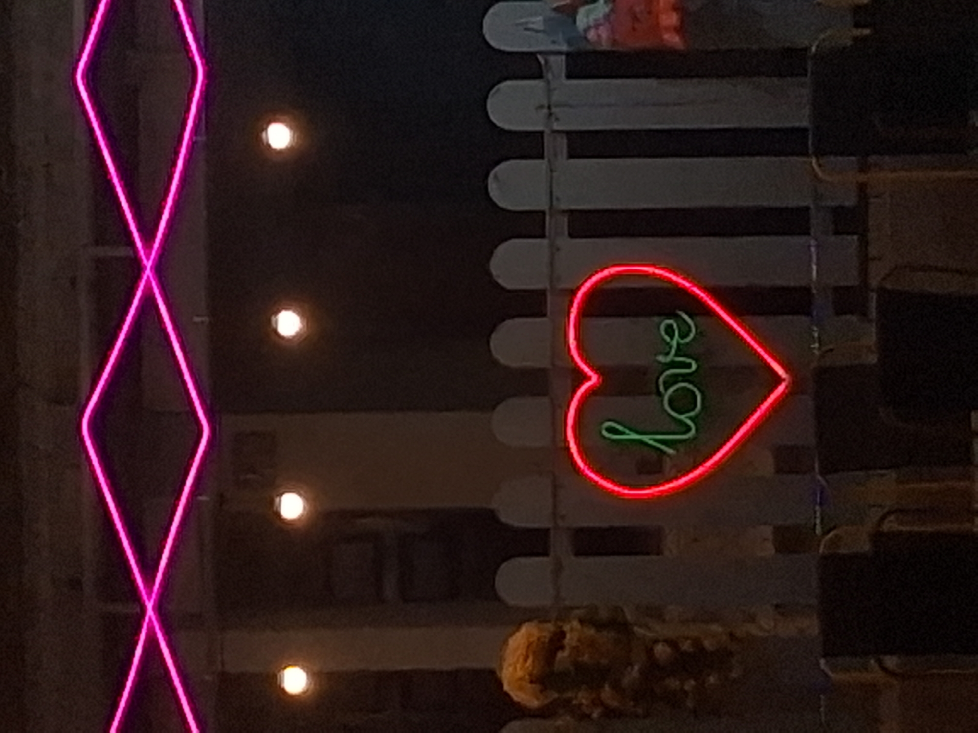 The image shows a neon heart with the word "love" written inside, glowing green, with a red outline, set against a dark background. Above the heart, pink neon lights form a zig-zag pattern. In the foreground, dark silhouettes of chairs are visible, suggesting an outdoor seating area. A white picket fence stands behind the neon heart. To the left of the heart, a portion of a textured, possibly decorative, object is visible. The overall scene appears to be in an urban setting at night, likely a cafe or restaurant in Phu Thuy Ward, Vietnam, given the context.