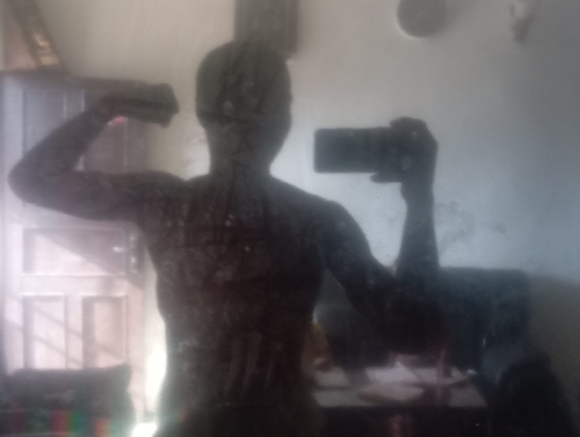 The photo is a blurry, low-quality image of a person taking a selfie in front of a mirror. The person is male, shirtless, and appears to be flexing their biceps. He is holding a smartphone in his right hand, presumably to take the photo. The background includes a window with bars and some indistinct objects. The location of Nkpor, Nigeria, cannot be confirmed or denied based on the image alone.