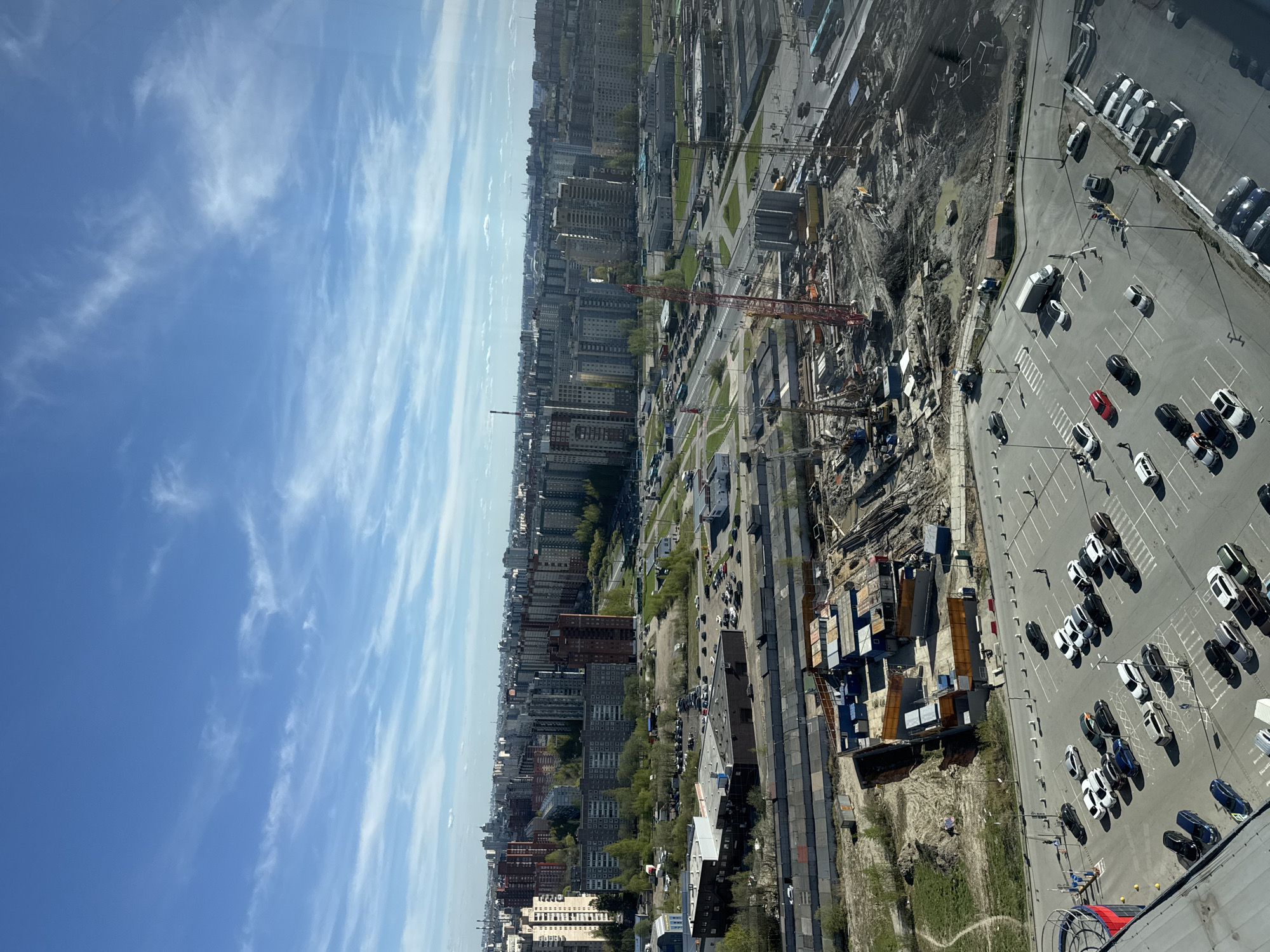 The image depicts an aerial view of a construction site and its surrounding urban area in Saint Petersburg, Russia.
A large, gray parking lot with numerous parked cars, mostly white, is visible to the right. Some vehicles are black or red.
Adjacent to the parking lot is a construction zone, characterized by excavated earth, heavy machinery, and building materials. A red construction crane is prominent within this area.
Beyond the construction site, a dense urban landscape unfolds with numerous tall residential buildings stretching into the distance. Trees are interspersed among the buildings.
The sky occupies a significant portion of the image, featuring a clear blue sky with elongated, wispy clouds.
