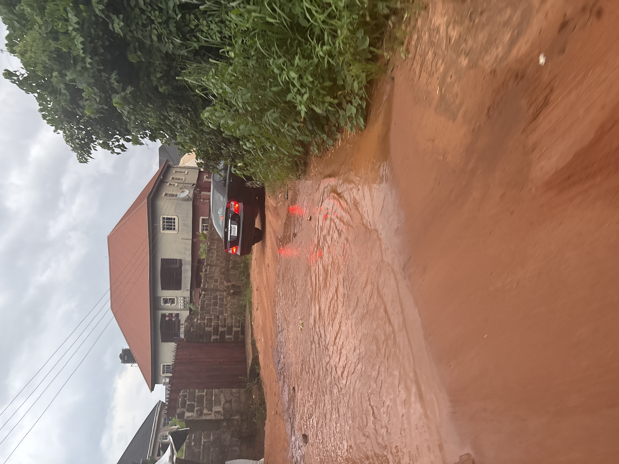 An overcast day in Obosi, Nigeria, shows a residential street after rainfall. A dark-colored sedan, with license plate NCW 319 SF, is parked on the red dirt road, its brake lights illuminated. Water pools significantly on the road, indicating flooding. To the left, a brick wall with a red gate encloses a property, beyond which a multi-story house with an orange roof is visible. To the right, dense green foliage lines the street. The scene depicts a common urban environmental condition in the area, likely a result of poor drainage exacerbated by recent precipitation.