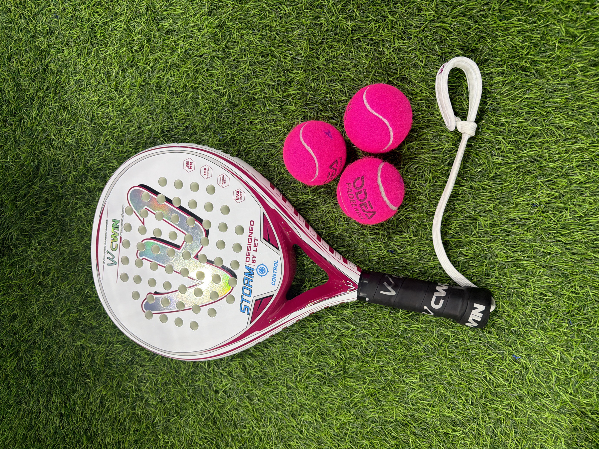 This media file shows a close-up, top-down view of a padel racket and three pink padel balls placed on artificial grass. The racket is white with pink and silver accents, featuring text that includes "WCWIN," "STORM," "DESIGNED BY LET," "CONTROL," "EVA SOFT," "36 MM," "TOP CONTROL," and "FRAME CARBON TECH." The balls are bright pink with white lines. A white wrist strap with a knot is attached to the end of the racket's handle, which is black and also has white text. The scene is brightly lit, suggesting an outdoor setting or a well-lit indoor space. There are no people visible, and the activity is implied to be related to the sport of padel. The artificial grass suggests a sports court environment. The image is static and provides no clues about the time of day or weather conditions, nor does it convey any specific emotions. The location context provided is Chaikovsky, Russia, but there are no visual cues within the image itself to confirm this.