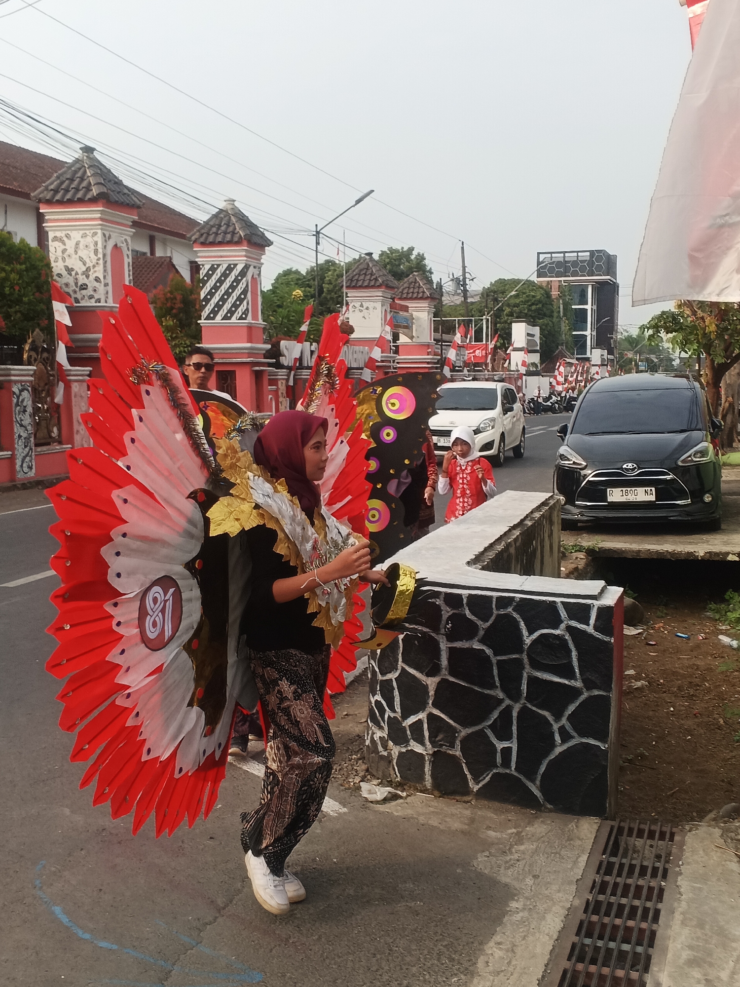 In Sokanegara, Indonesia, a parade is underway on a paved road lined with decorative pillars and flags. A young woman, dressed in a hijab and a traditional patterned outfit, is the central figure, adorned with a large, vibrant red and white wing-like costume with gold accents. The costume features a circular emblem with the number "81." To her right, another child, also in traditional attire, walks in the parade. Behind the woman, a man wearing sunglasses watches the event. Several vehicles, including a black Toyota Sienta and a white car, are visible on the road, some parked and others in motion. The environment appears to be a street scene with buildings, trees, and overhead power lines under an overcast sky.