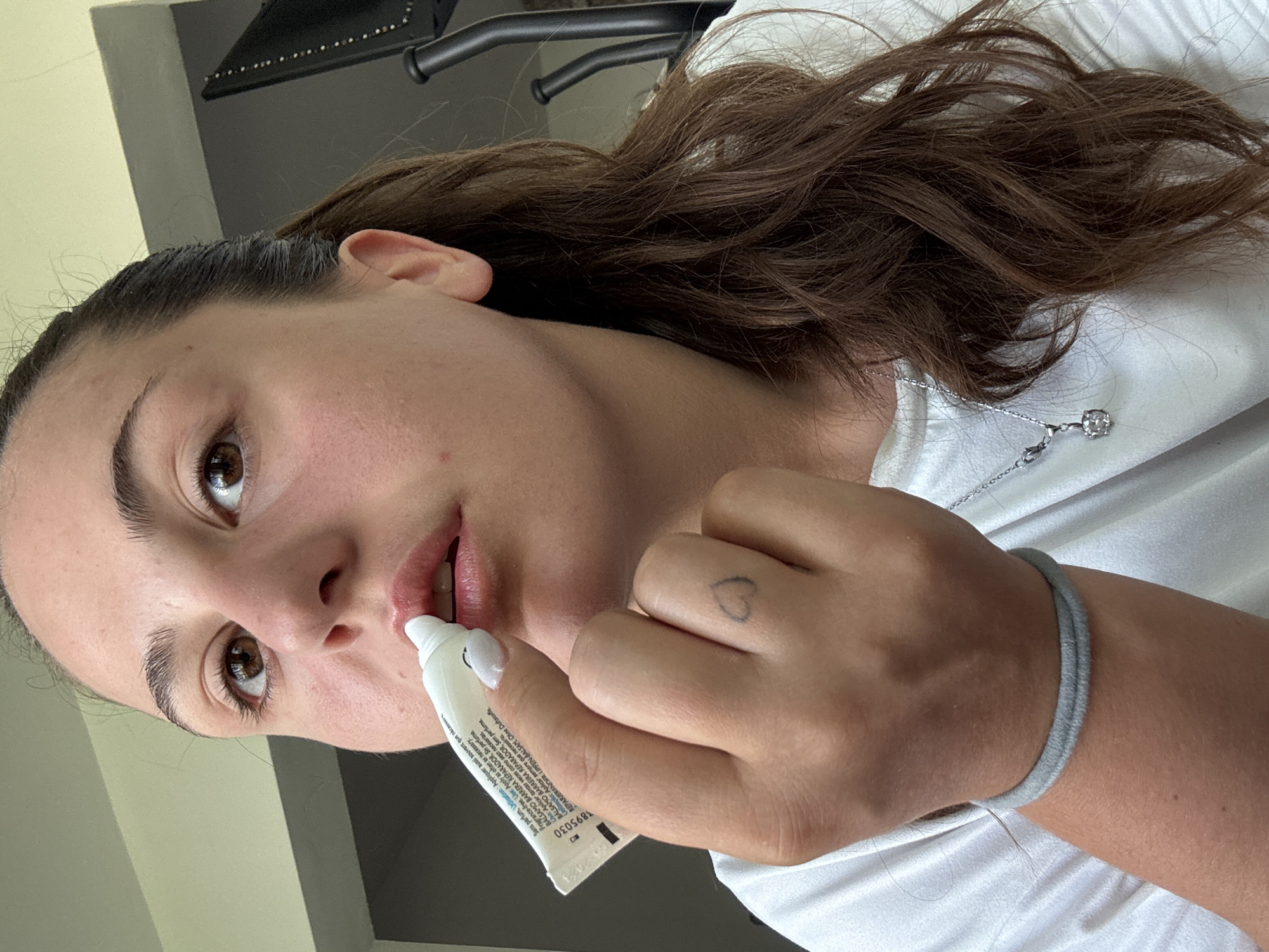 A young woman is applying lip balm to her lips. She is holding a white tube of lip balm in her right hand, and the applicator tip is touching her lower lip. Her eyes are brown and she has dark brown hair pulled back in a ponytail. She is wearing a white t-shirt, a grey bracelet on her left wrist, and a silver necklace with a pendant. A small heart tattoo is visible on the back of her right hand. The background is out of focus, but appears to be an indoor setting with some exercise equipment. The image was taken in Plovdiv, Bulgaria.