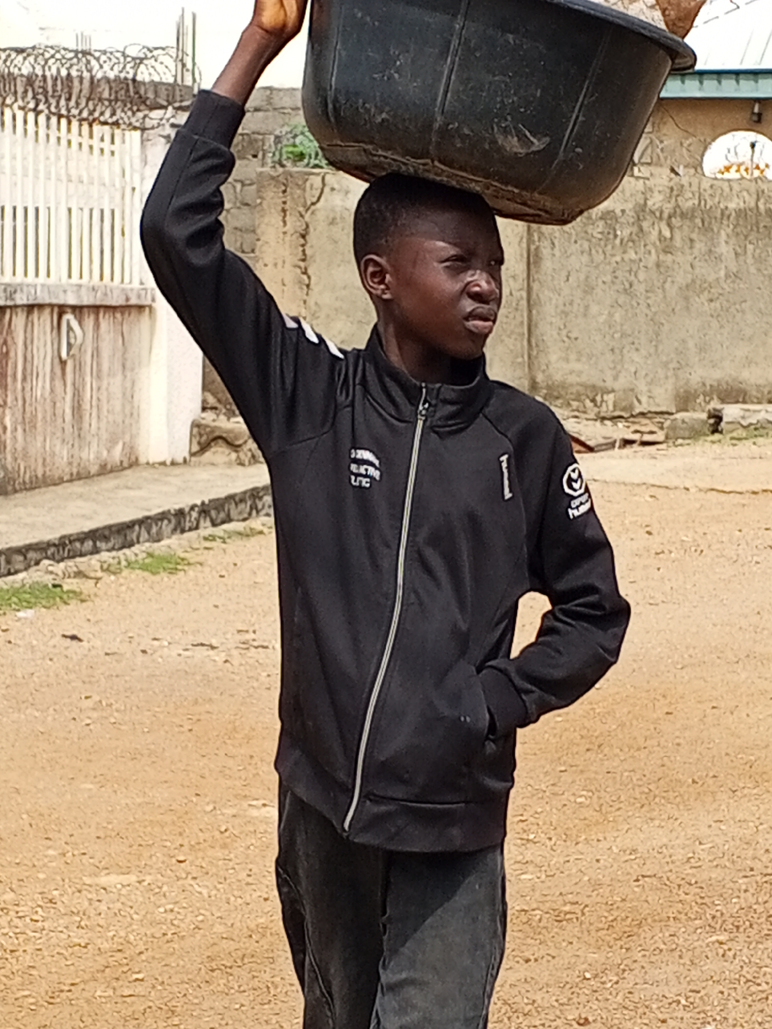 The image displays a young boy, likely of African descent, carrying a large, dark-colored plastic basin or tub on his head. He uses his raised right arm to stabilize the load. The boy is positioned centrally, facing slightly to his right, with a concentrated or serious expression.
He is dressed in a black long-sleeved tracksuit jacket with a full-length zipper and dark pants. The jacket features white stripes on the upper arms. On the left chest, there is a vertical white design resembling the letter "I" or the number "1," with text "GWAZ ACTIVE LTC" visible below it. On the right chest, a white circular logo is present, with the words "SPORT HUMMEL" partially legible beneath it.
The setting appears to be an outdoor, unpaved area with a light brown, sandy or dirt ground. In the background, there are light-colored walls of buildings, and to the left, a white slatted fence topped with barbed wire is visible. The bright, even lighting suggests it is daytime. The scene depicts a common method of transporting goods, consistent with a semi-urban or rural environment like Adankolo, Nigeria.