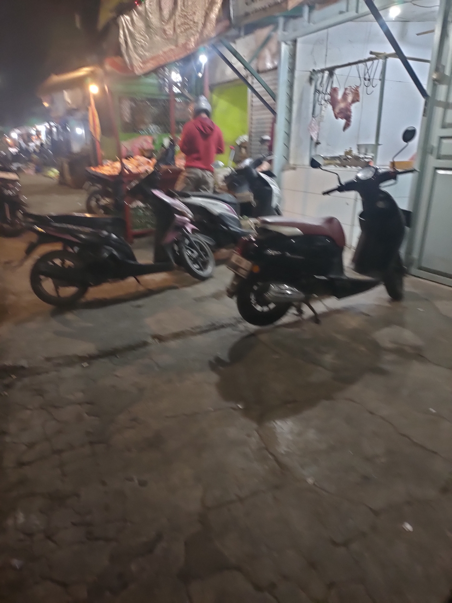The image depicts an outdoor scene at night in a market area, possibly in Subang, Indonesia. Several motorcycles are parked on a paved surface, some with their headlights illuminated, casting a warm glow. In the background, a person wearing a red hoodie stands near a stall with a tarp overhead, suggesting a food or goods vendor. The environment appears to be a bustling marketplace, with glimpses of other stalls and structures. Meat is hanging from a rack in the upper right portion of the frame, indicating a butcher's stall or a similar establishment. The overall atmosphere is lively and characteristic of a night market.