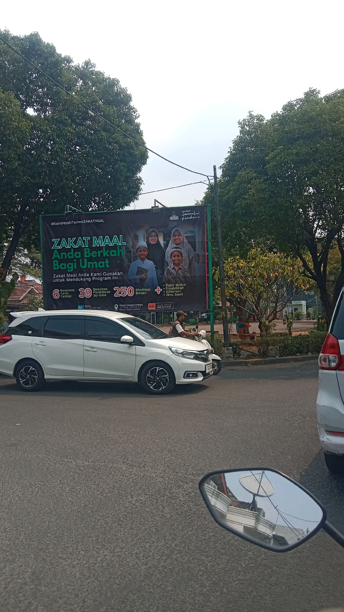 The image shows a street scene in Bekasi, Indonesia, with a large banner displayed on the left. The banner, which is black and green, advertises "ZAKAT MAAL Anda Berkah Bagi Umat," meaning "Your Zakat is a Blessing for the Ummah." It features images of two women and two boys, all wearing Islamic attire. Text below indicates that Zakat Maal is used to fund programs. Numbers 6, 39, and 250 are prominent, associated with "Beasiswa Santri Tahfidz" (6 scholarships for memorizers of the Quran), "Beasiswa Pendidikan SD-SMA" (39 scholarships for primary and high school education), and "Khidimat Binaan" (250 services/assistance). A list of recipients of zakat includes "Fakir Miskin" (the poor), "Fisabilillah" (those fighting for a cause), "Ghorimin" (debtors), and "Ibn Sabil" (travelers). Contact information for "Masjid Jamiatul Fadhus" is also present.
In the foreground, a white Honda Mobilio is parked, and a white motorcycle with a rider is moving past it. The rider appears to be wearing a brown shirt and a helmet. The street is paved, and there are trees on the left and right sides of the banner, suggesting an outdoor setting. In the reflection of the motorcycle's side mirror, a satellite dish and some buildings are visible. The sky is overcast and light. The scene suggests a busy urban environment in Bekasi, Indonesia.