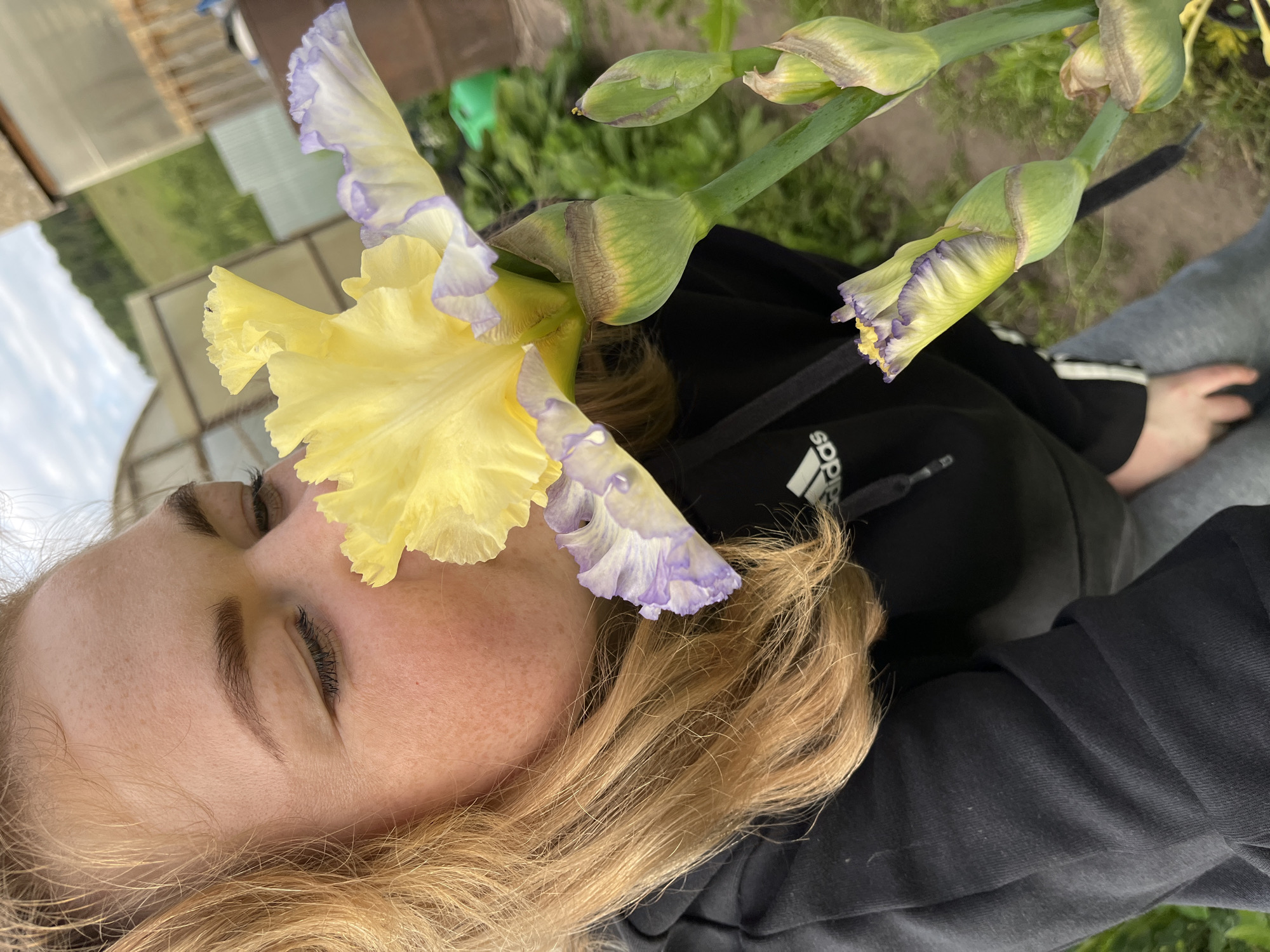 A young Caucasian female with blonde hair is seen in profile, smelling a yellow and purple iris flower. She is wearing a black hooded sweatshirt and grey sweatpants. The background suggests an outdoor setting, possibly a garden or rural area, with greenery, a greenhouse structure, and a barrel visible. The sky appears overcast. The location is identified as Olkhovka, Russia.