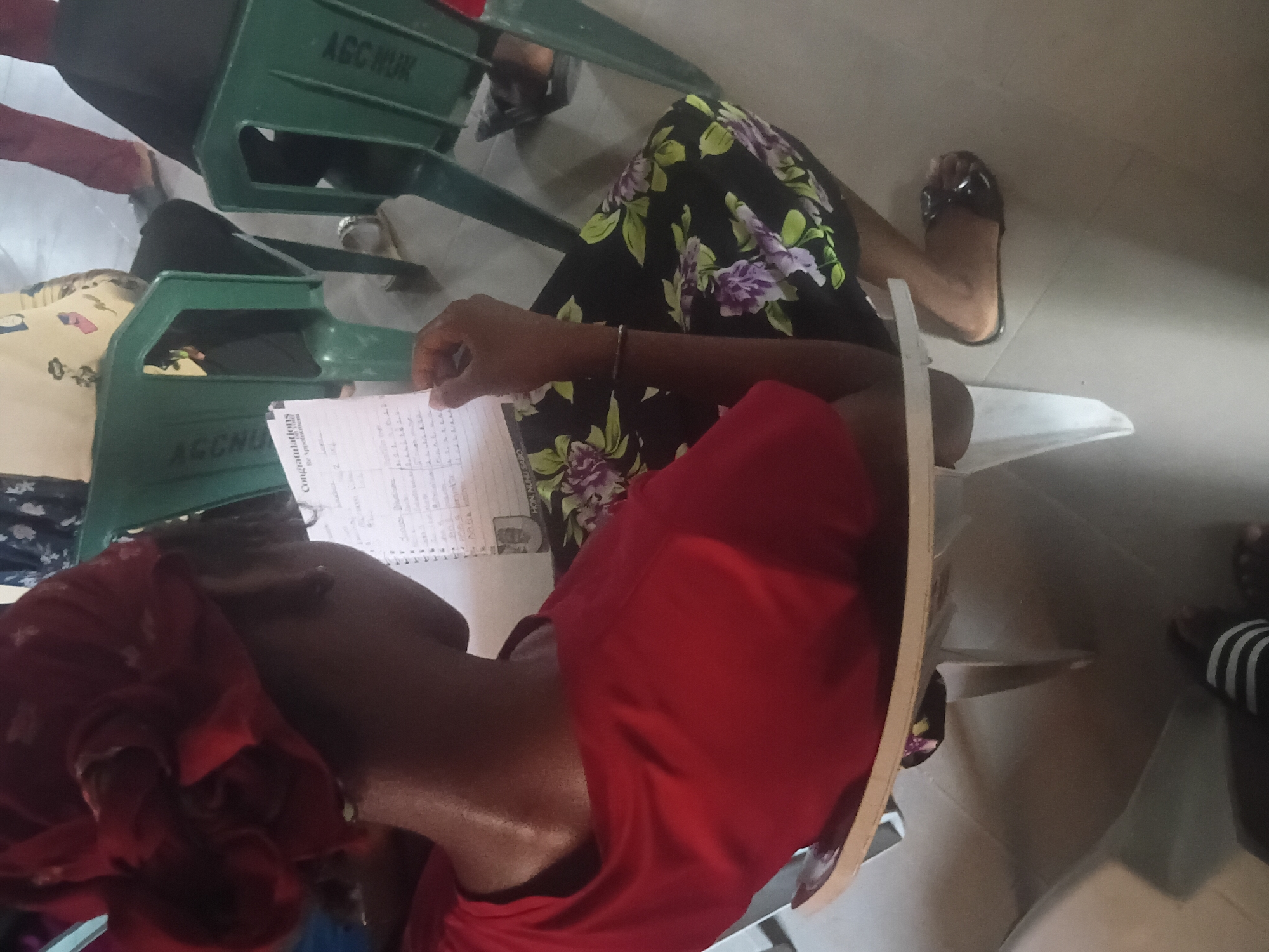 In the city of Jalingo, Nigeria, an indoor scene depicts an individual seated on a light-colored chair, viewed from behind and slightly to the right. The person, appearing to be an adult female with dark skin, wears a reddish-orange fabric headwrap, a red short-sleeved top, and a black skirt with a pattern of green leaves and purple flowers. A thin, dark bracelet is on her right wrist, and she wears a dark slide sandal on her right foot. She holds an open notebook or a piece of paper in her hands, with visible handwritten text including "Congratulations" and "for Appointment" at the top, followed by lists of names or items. In the background, two green plastic chairs are visible, each bearing the inscription "AGCNUR" on its backrest. Other individuals are partially visible in the background, including legs and feet, one pair wearing maroon pants and black shoes, and another pair wearing striped slide sandals in the lower-left foreground. The floor is composed of light-colored tiles.