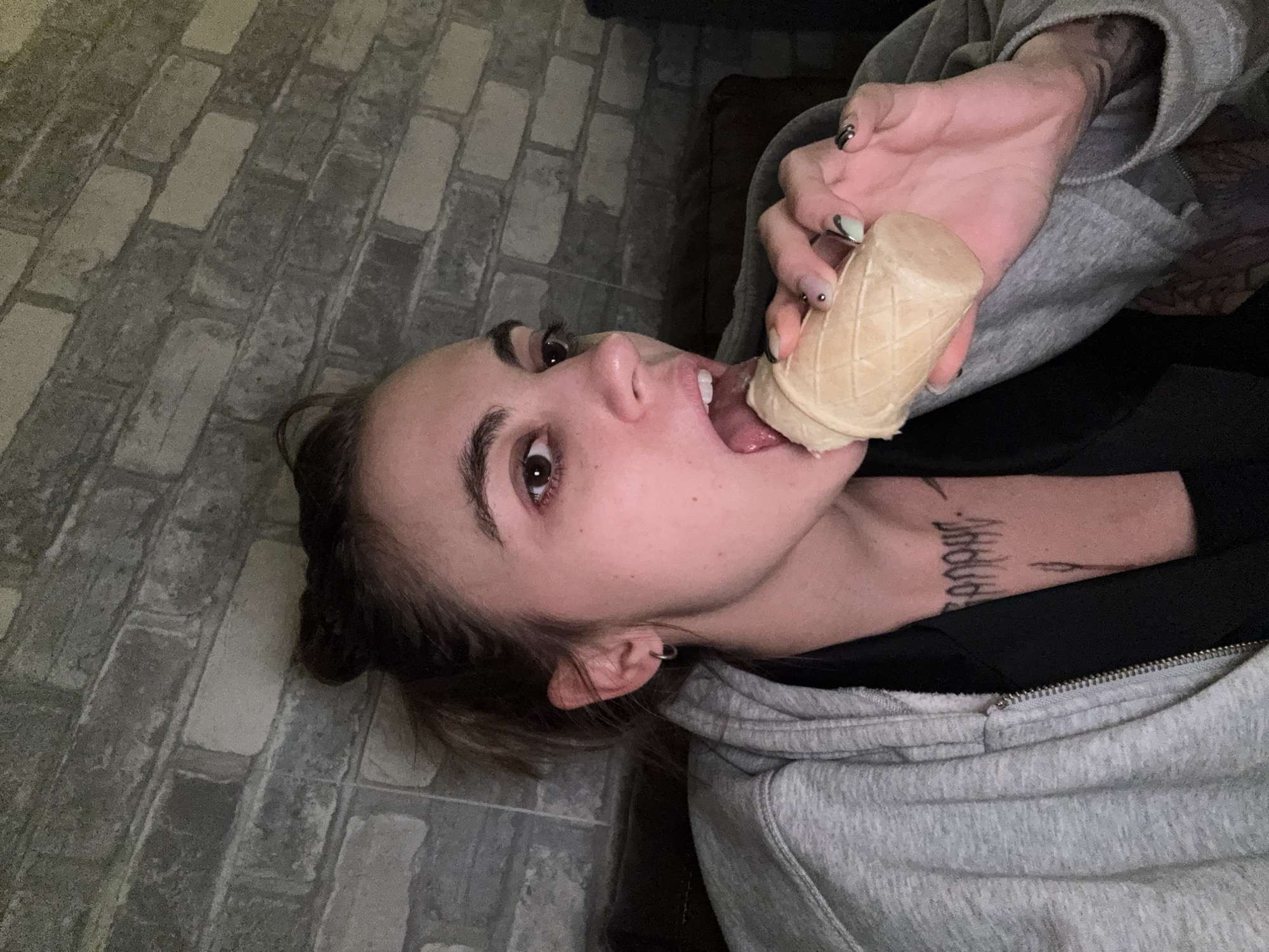 A young woman with dark hair is captured in a close-up shot, lying on her side. She is wearing a grey hooded sweatshirt and has a tattoo on her neck. In her right hand, she holds an ice cream cone, and her tongue is extended towards it, suggesting she is about to eat the ice cream. Her eyes are wide and looking directly at the camera, and her expression seems playful or provocative. The background appears to be a textured wall, possibly made of brick or a similar material, suggesting an indoor setting. The lighting is dim, hinting that it might be nighttime or a dimly lit room.