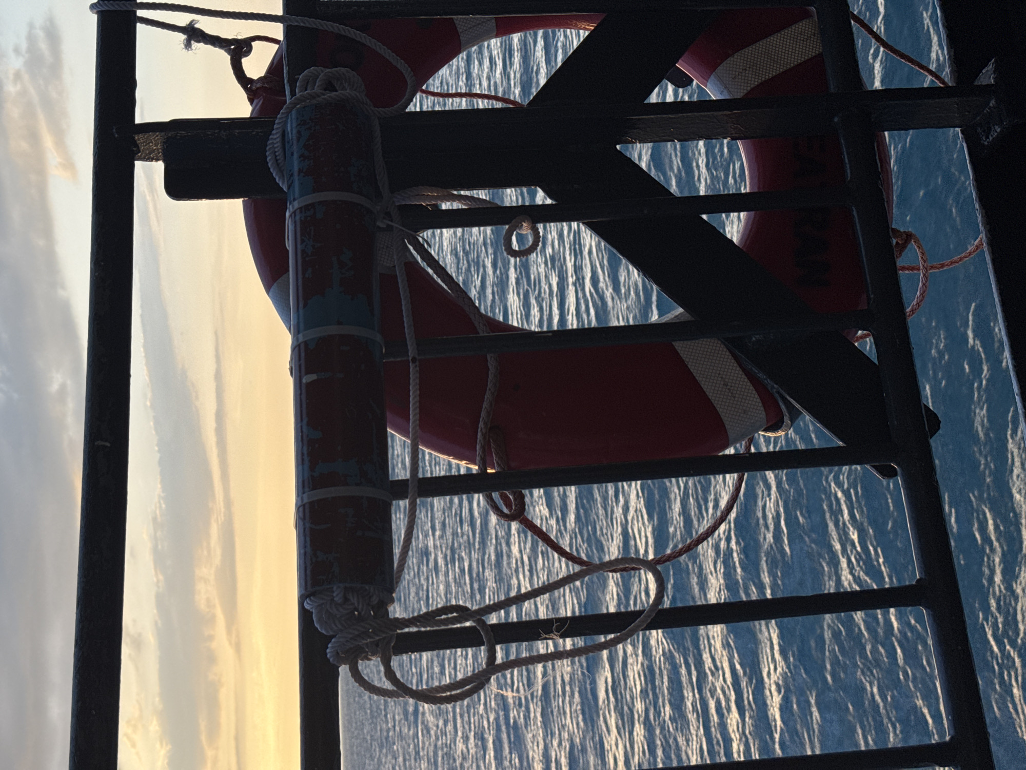 The image shows a close-up, eye-level view of a life preserver attached to a railing, with the ocean and a cloudy sky in the background. The life preserver is red and white, with some blue and white markings on its surface. It is secured by ropes. The railing is made of black metal. The ocean is calm and appears to be a deep blue, with sunlight reflecting off the surface. The sky is filled with light clouds, and the sun appears to be setting or rising, casting a warm glow. The scene suggests a maritime setting, possibly on a ferry or a ship. The location is Unknown, Thailand. There are no people visible in the image.