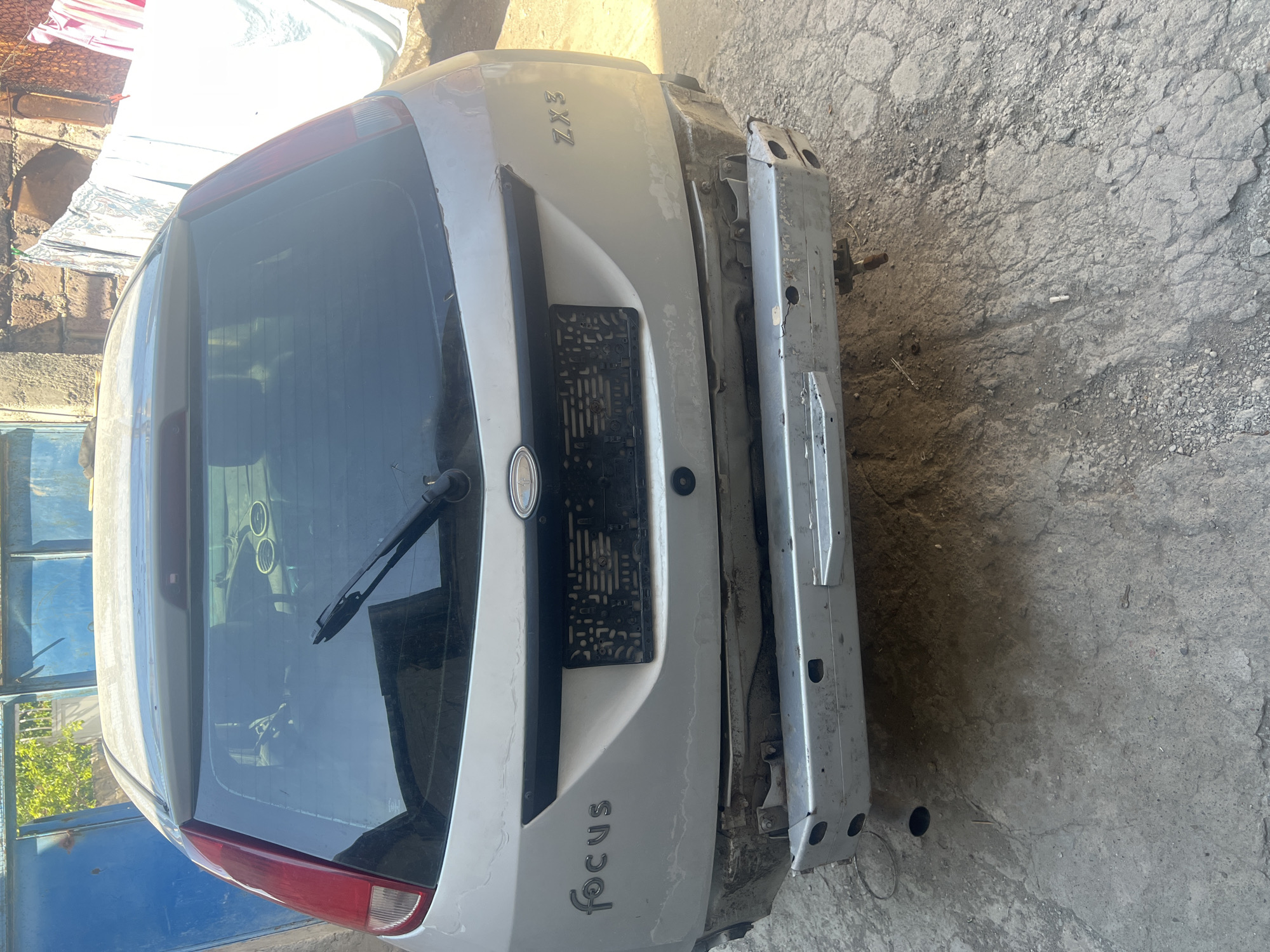 An unoccupied, silver Ford Focus ZX3 hatchback, is parked outside on a dirt and concrete surface. The car is viewed from the rear, and the bumper is missing, exposing the car's frame and metallic components. The car's taillights and rear window are visible, as is a windshield wiper. The background shows a brick wall, blue garage doors, and laundry hanging on a clothesline. The sky is bright, indicating a sunny day.
