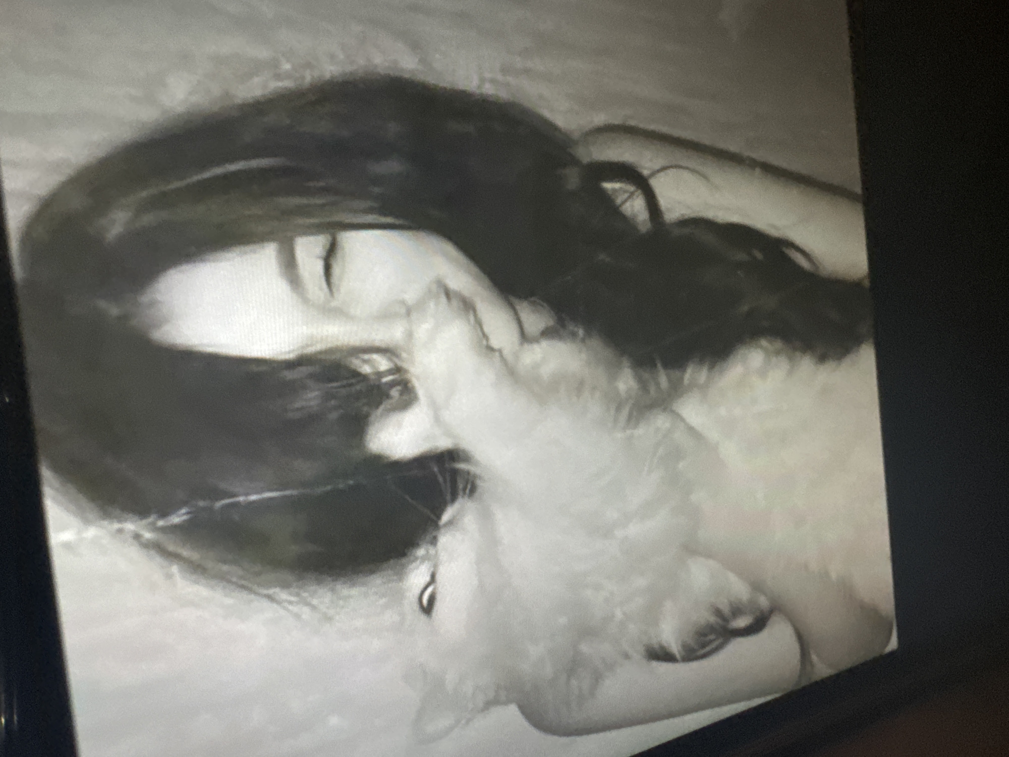 A monochrome image depicts a person with long, dark hair lying down, engaged in a tender interaction with a small, fluffy animal. The person's eyes appear closed or mostly closed, suggesting a moment of rest, affection, or sleep. Their face is partially obscured by the animal, which is nestled closely against their cheek and mouth area. The animal has light-colored, voluminous fur and a dark snout, likely a small dog or a cat.
The scene takes place indoors, with a light, plain background, possibly bedding or a wall. The lighting is dim, contributing to an intimate and peaceful atmosphere. The image has a soft-focused or slightly grainy quality, enhancing its artistic and personal feel. The perspective is slightly angled from above. No text is visible in the image. The overall mood conveys warmth, companionship, and a quiet moment shared between the individual and their pet.