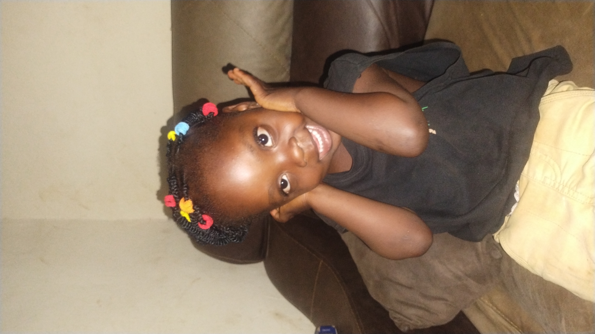 The image displays a young, dark-skinned child, estimated to be of pre-school age, seated and centrally positioned, facing the viewer directly. The child's hair is styled in numerous small braids or twists, accented with multi-colored elastic bands or beads in red, yellow, and blue. She is dressed in a dark, likely black, short-sleeved t-shirt and light brown or khaki-colored shorts. Her arms are bent, with her hands placed near her cheeks/ears in a posed manner. She exhibits an open-mouthed smile, revealing teeth, and maintains direct eye contact. The child is seated on a brown, upholstered piece of furniture, consistent with a sofa or chair, with a lighter brown cushion visible to her left. The background comprises plain, light-colored walls, appearing beige or off-white, with an interior corner visible in the upper left portion of the frame. The scene is indoors, indicative of a residential or domestic environment. The image contains no discernible visual information, such as landmarks, specific architecture, or textual indicators, to confirm the location as the city of Jos, Nigeria.