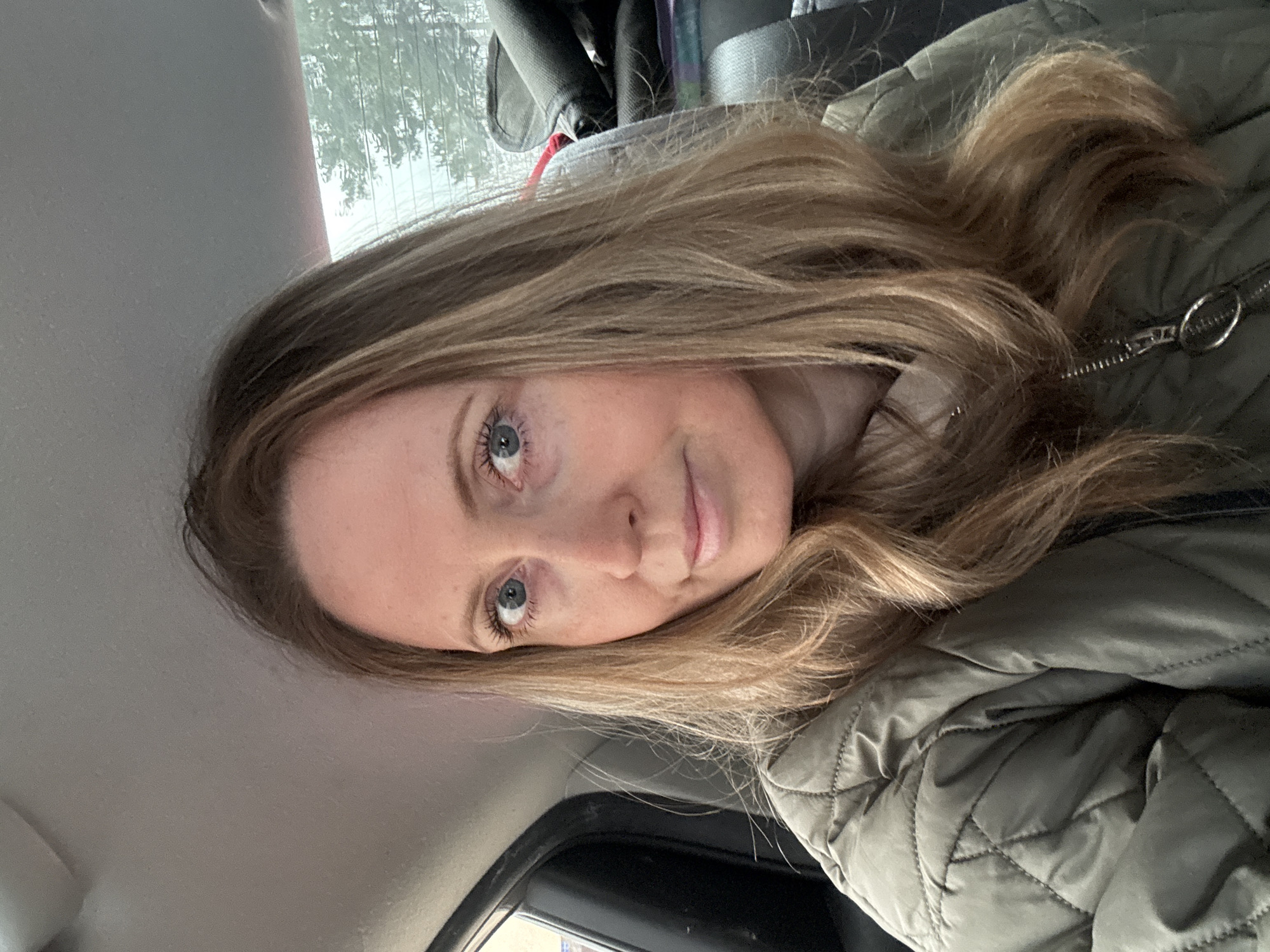 The image shows a woman with long blonde hair and blue eyes. She is wearing a quilted green jacket. The background suggests she is inside a vehicle, likely a car, with a window visible. The location in Chaikovsky, Russia, cannot be determined from this image alone. There are no specific landmarks, signs, or other indicators to identify the location. The scene primarily depicts the woman in the car.