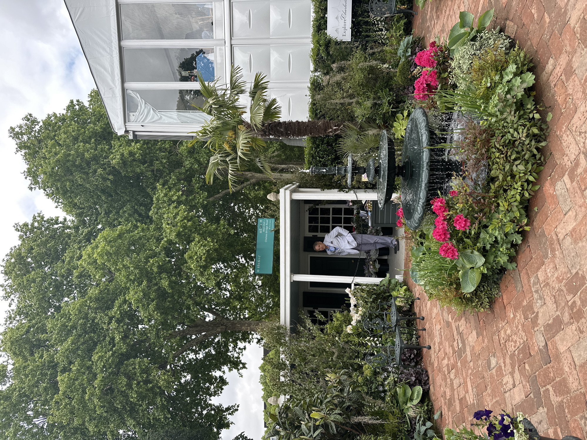 A verdant outdoor setting is displayed, featuring brick paving, manicured vegetation, and a white gazebo structure. A multi-tiered fountain sits amid a garden of flowering plants with pink blossoms. A person stands under the shelter of the gazebo, while a palm tree punctuates the scenery nearby. A building with prominent windows is seen to the left, with its details visible. The scene is located in London, United Kingdom.