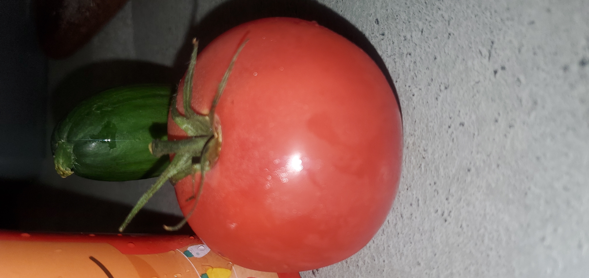 The image shows a close-up of a ripe, red tomato with water droplets on its surface, resting on a speckled gray countertop. To the left, a portion of an orange container with a smiley face design is visible, also with water droplets. Behind the tomato, a green cucumber is partially visible, its skin exhibiting vertical ridges. The lighting suggests an indoor setting, casting shadows on the countertop. No specific location in Sasbach, Germany can be determined from the visual information alone.