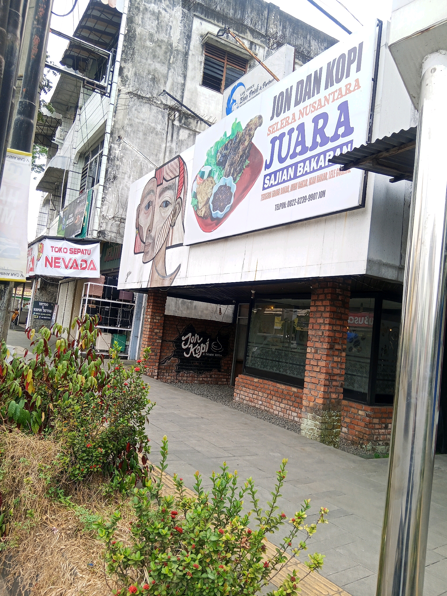 The image depicts the facade of a commercial building in Lubuklinggau, Indonesia. On the upper level, a large white banner advertises "JON DAN KOPI SELERA NUSANTARA JUARA SAJIAN BAKARAN" which translates to "Jon and Coffee Nusantara Taste Champion Grilled Dishes." Below the banner, a stylized mural of a human face is visible. Adjacent to this, a smaller banner reads "TOKO SEPATU NEVADA" indicating a shoe store. The ground floor features a brick-faced establishment with large glass windows, with a sign reading "Jon Kopi & Seafood" visible inside. The street level is paved with gray tiles and bordered by a yellow tactile paving strip. Green bushes with red flowers are planted in front of the building, and a dry, brown grassy area is also present. The overall scene conveys a commercial streetscape with various businesses.