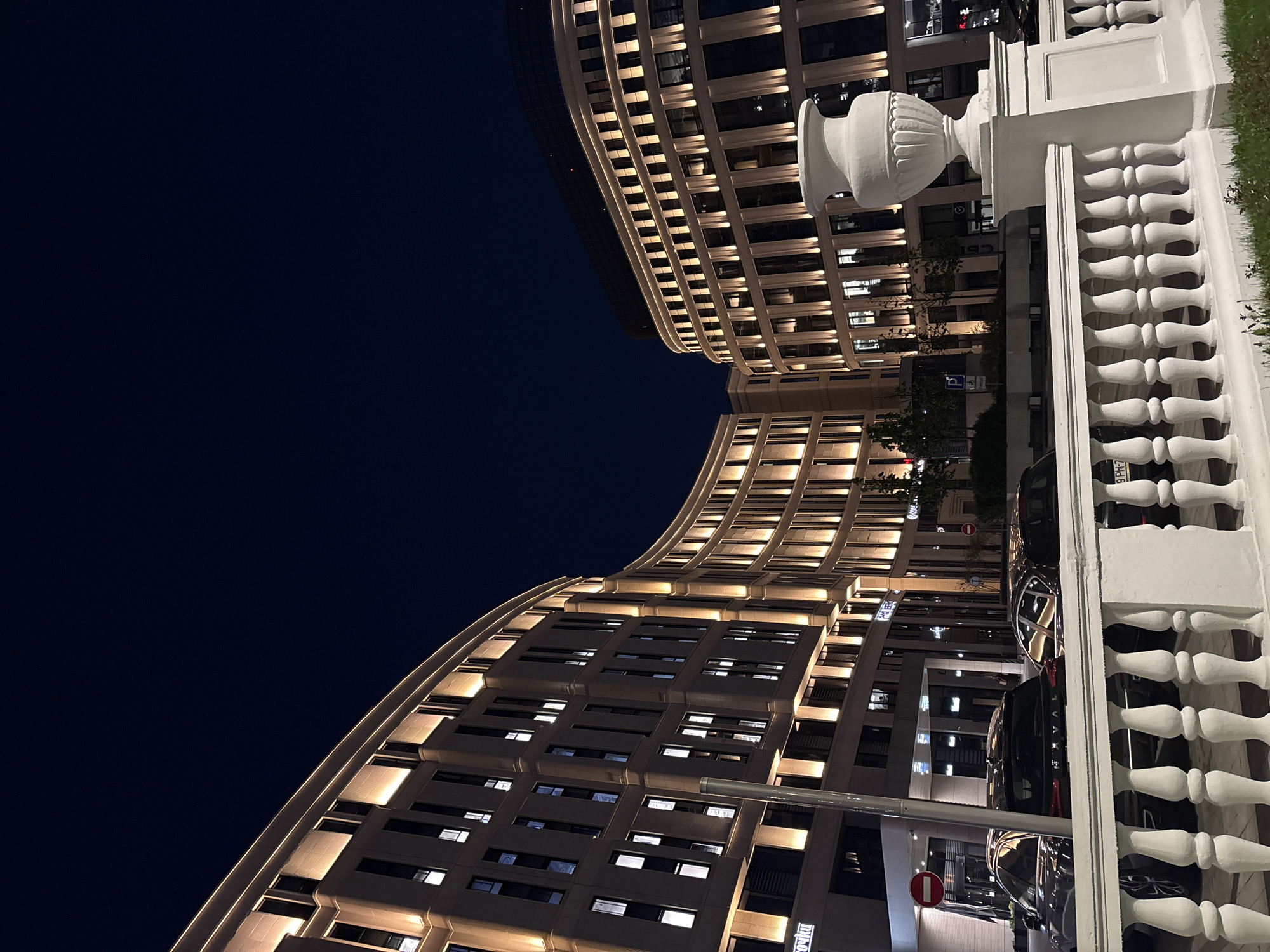 The photograph depicts a night view of an architecturally distinctive building in Minsk, Belarus. The structure exhibits a curved facade with illuminated windows, creating a grid-like pattern against the dark night sky. A white balustrade with a decorative urn is prominently featured in the foreground, partially obscuring a row of parked cars. A street sign is visible near the building's base, indicating a controlled traffic area. The sky is a deep blue, suggesting a late evening or nighttime setting.