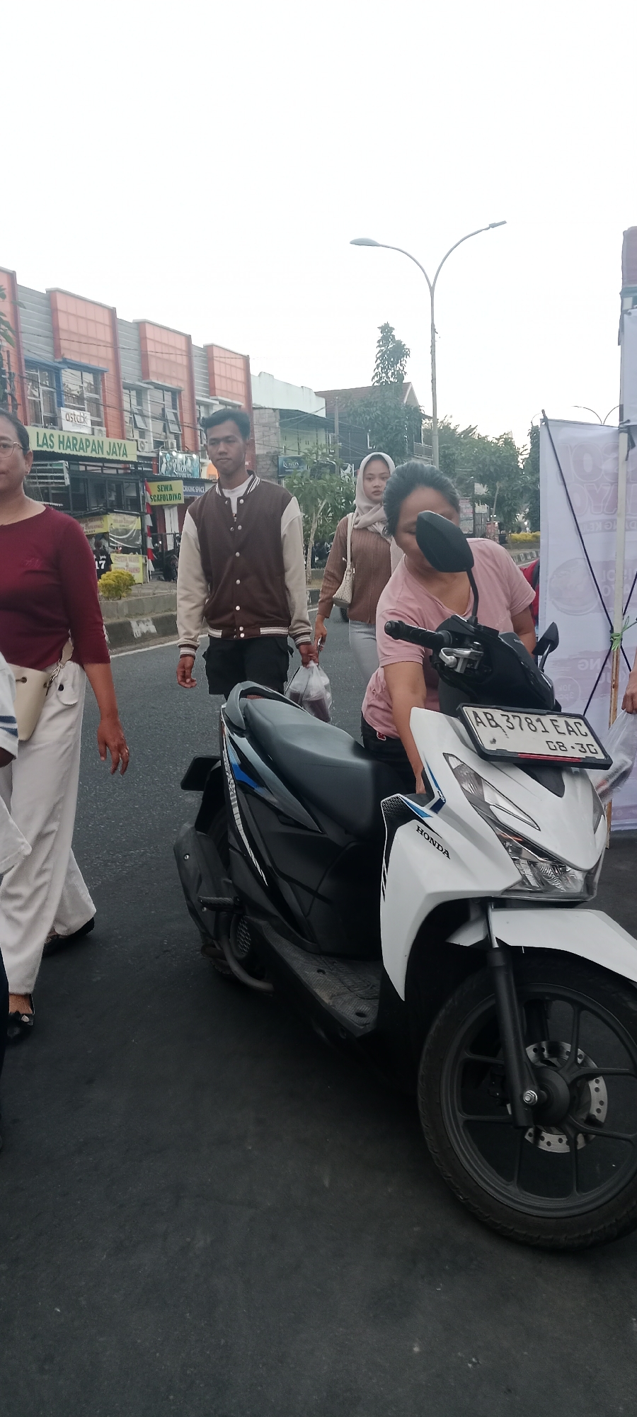 In Ngemplak, Indonesia, a woman wearing a pink t-shirt is seated on a white and black Honda scooter, with her right hand on the handlebar and her head bent down towards the scooter. She is positioned on the right side of the frame. To her left, a man in a brown and beige jacket and black shorts walks away from the camera, carrying a plastic bag in his right hand. Behind him, a woman wearing a light brown hijab and a beige sweater walks with a purse slung over her shoulder. Further to the left, partially visible, is a woman in a maroon shirt and white pants. The scooter's license plate reads "AB 3781 EAC 08-30". The scene takes place on a paved street with buildings and trees in the background. The sky is overcast.