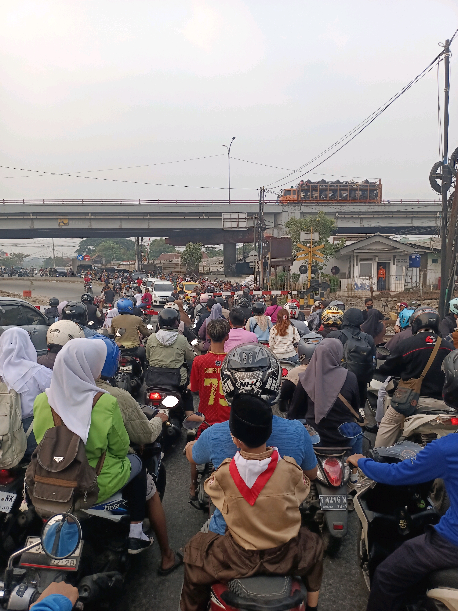 In Karawang, Indonesia, a dense traffic jam occurs at a railroad crossing. A multitude of motorcycles and a few cars are stationary on the asphalt road. The sky is overcast, and a pedestrian overpass spans the road in the background. A truck carrying goods is visible on the overpass. A railway signal with yellow crossbars indicates that the crossing is active, and a small building sits beside the tracks. Several individuals are riding motorcycles, with many wearing helmets and headscarves. One person in the foreground is wearing a scout uniform. The scene captures a typical urban traffic situation in Indonesia.