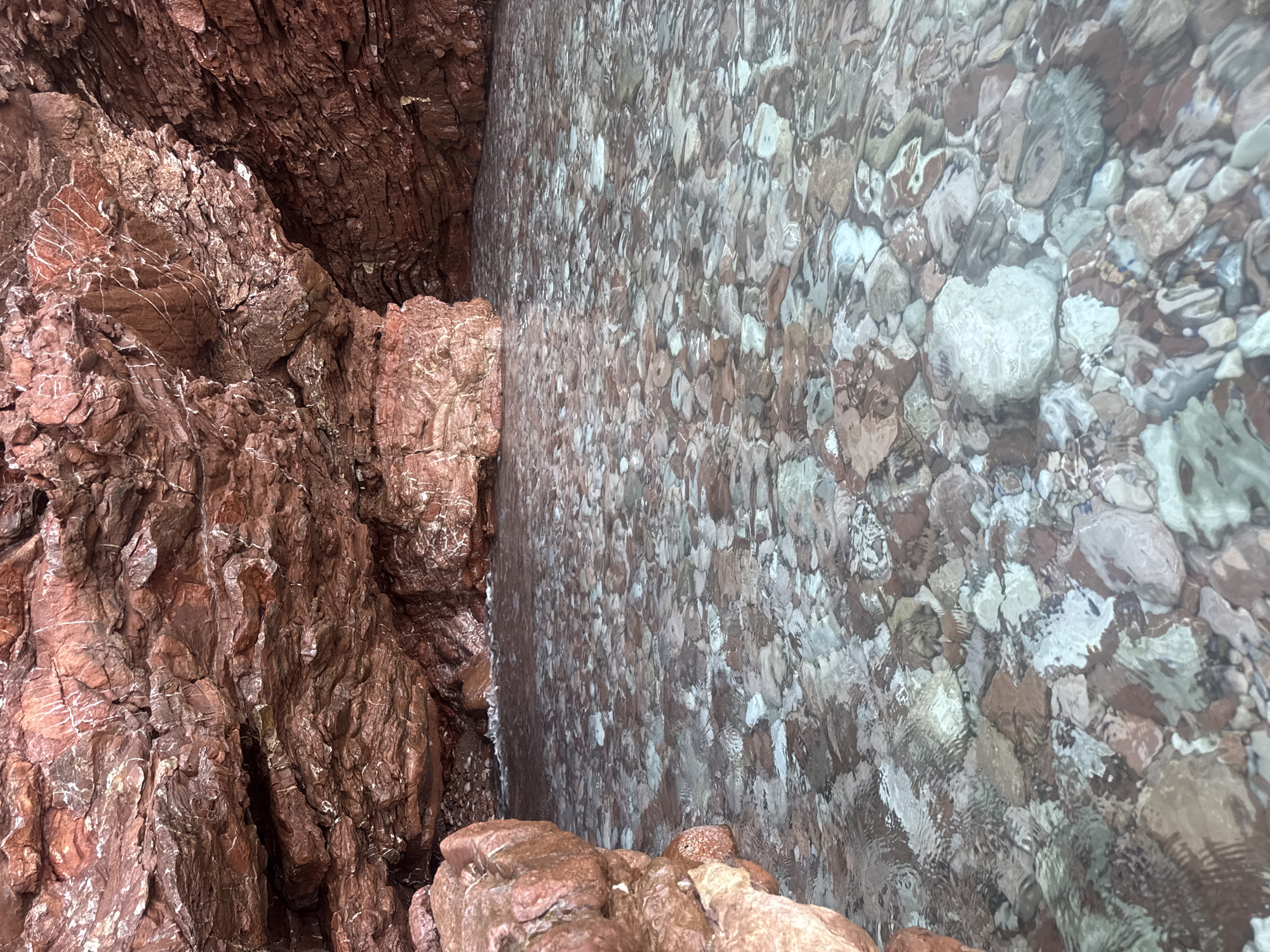 The image shows a close-up view of a rocky shoreline with clear, shallow water. On the left, rugged, reddish-brown rocks are piled and layered, showing intricate textures and striations, with some lighter, white veins running through them. A narrow gap between these rocks leads to a darker recess. To the right of the rocks, the water covers a bed of smooth, rounded pebbles and stones. The stones are varied in color, including shades of brown, grey, white, and green, and their shapes are softened and distorted by the rippling surface of the water. The water is transparent enough to reveal the details of the stones beneath, but the movement of the water creates a shimmering, mosaic-like effect.
The scene appears to be an outdoor natural environment, likely a coastal area. The lighting suggests it could be daytime, with the sun illuminating the rocks and reflecting off the water. There are no people or specific objects visible, only the natural elements of rock and water. The overall impression is one of a serene and natural coastal landscape, characteristic of a Mediterranean or similar climate. The context provided, Sveti Stefan, Montenegro, suggests a location known for its picturesque coastline and clear waters. There is no visible text in the image.