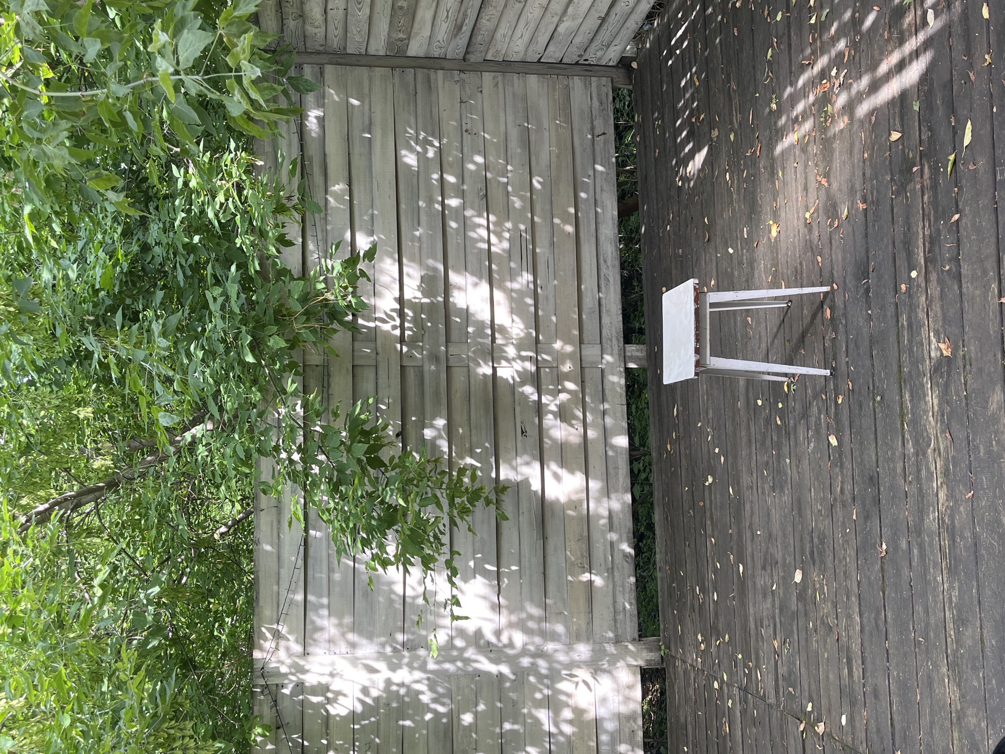 The image shows an outdoor scene with a wooden deck and a lighter-colored wooden fence. On the left side of the frame, lush green foliage from a tree hangs over the fence, casting dappled shadows on its surface. The wooden fence is made of vertical planks and appears weathered.
On the right side of the frame, the wooden deck is made of darker, horizontal planks. The deck is scattered with fallen leaves, suggesting an autumn or late summer setting. Sunlight filters through the leaves of an unseen tree, creating intricate patterns of light and shadow on both the fence and the deck.
A small, white, metal stool with a simple design is placed on the wooden deck. The stool has a rectangular seat and four thin legs. The scene is illuminated by natural sunlight, and the shadows suggest it is daytime. The overall impression is one of a quiet, natural outdoor space, possibly a backyard or garden area. There is no visible text in the image.
