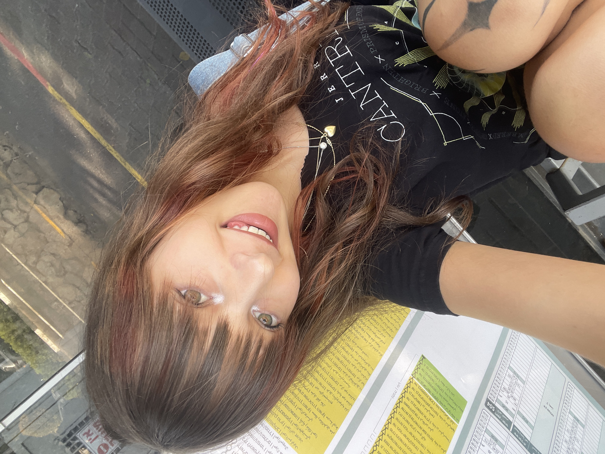 A young woman with long, brown hair, streaked with pink, smiles at the camera. She is wearing a black t-shirt with a graphic design and a denim jacket. She is adorned with a gold heart-shaped pendant necklace. The woman is sitting outdoors, with what appears to be a bus schedule or informational display board to her left. In the background, a street with yellow and red lines is visible, along with some foliage. The scene suggests she might be at a bus stop or waiting area in the city of Raanana, Israel.