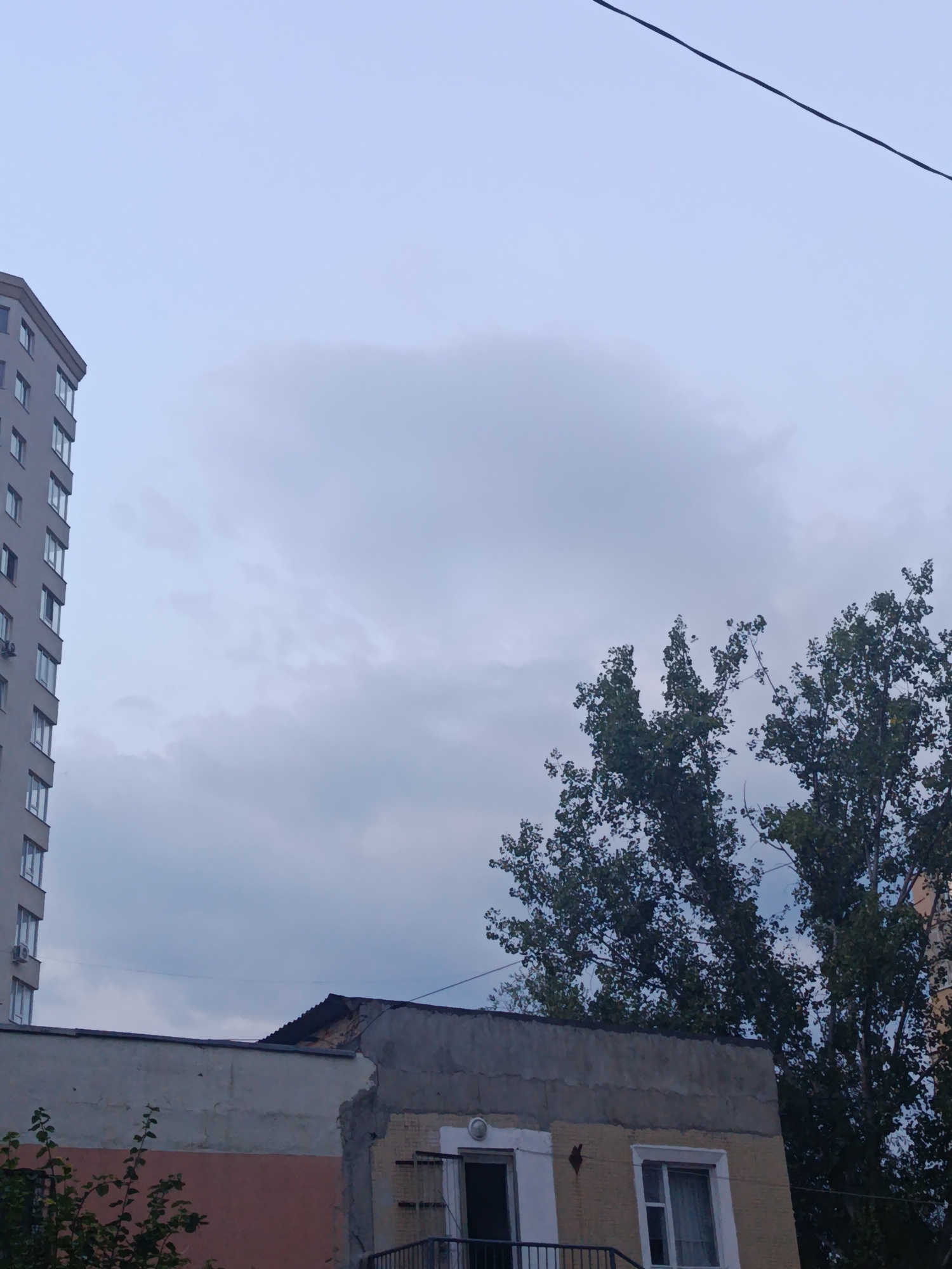 This image captures an overcast sky over a cityscape, likely in Chișinău, Moldova, given the typical architectural style. On the left, a tall, modern apartment building with a neutral gray facade and numerous windows stands prominently. To the right of the apartment building, a lower, older structure is visible, featuring a partially damaged or unfinished roof with visible corrugated metal. Trees with dense green foliage partially obscure the view of these buildings, suggesting a late spring or summer season. A dark, thin wire stretches diagonally across the upper portion of the frame, indicating the presence of overhead electrical or communication lines. The overall impression is one of urban infrastructure under a muted sky. No people or specific actions are depicted.