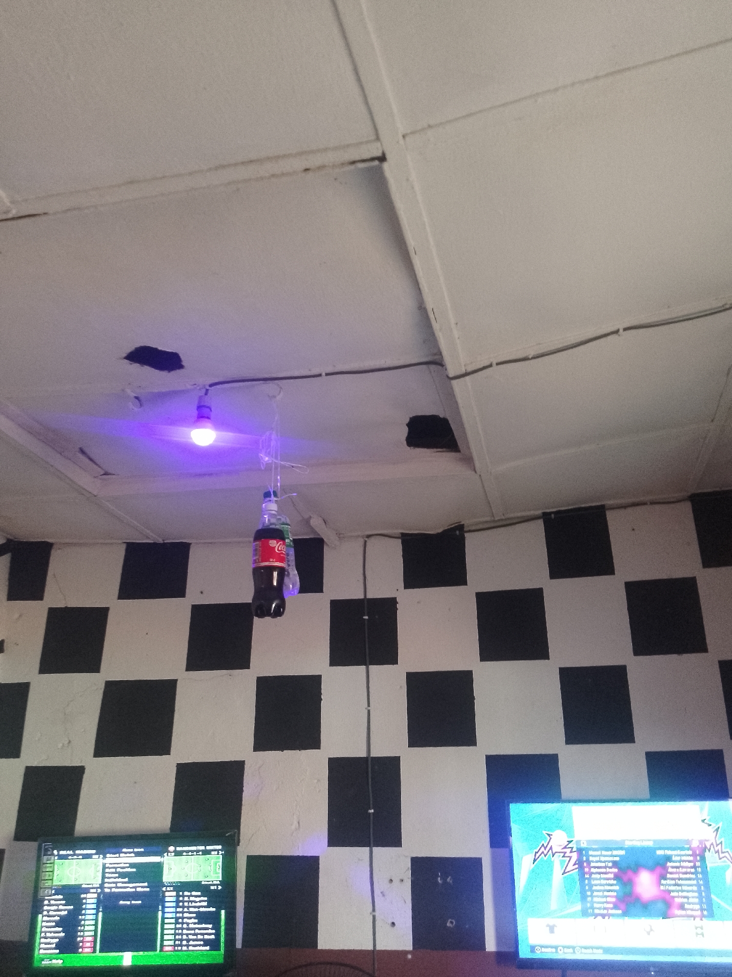 In Serti, Nigeria, a dimly lit room features a checkered black and white wall. A single fluorescent bulb emits a purple glow, illuminating a Coca-Cola bottle hanging by string from the ceiling. Two large televisions are visible, one displaying a sports game with team lineups and player statistics, and the other showing a vibrant, abstract graphic. The ceiling is made of white panels, some of which are damaged, revealing dark openings. Electrical wires are visible along the ceiling.