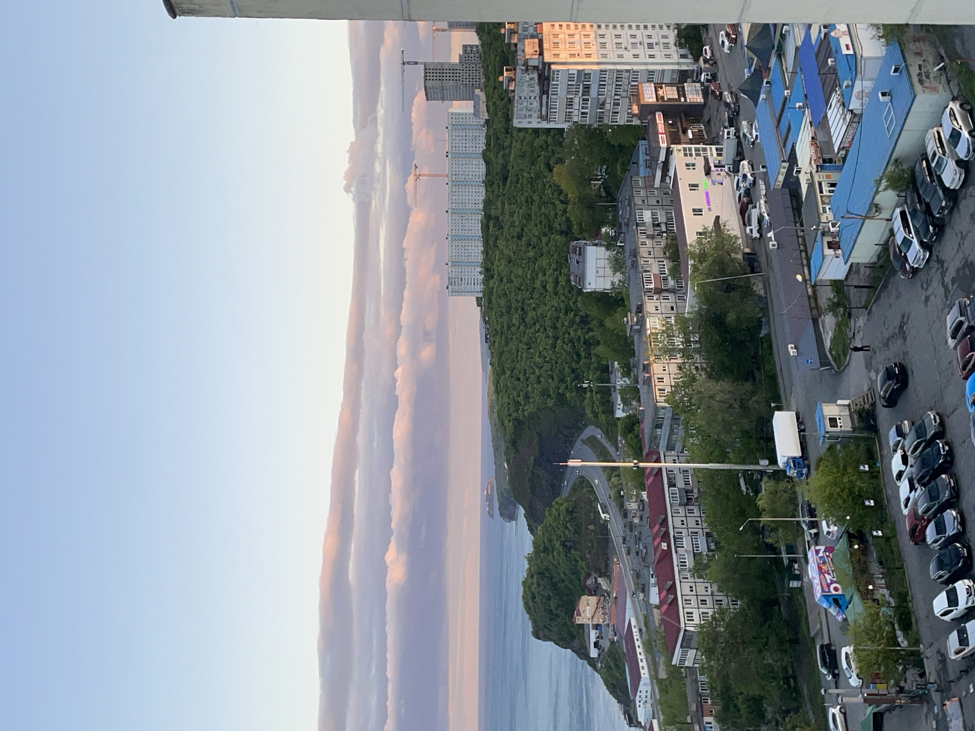 The image presents an elevated view of Vladivostok, Russia, likely taken from a high-rise building. The scene is divided into several distinct areas: a sky with pastel-colored clouds, a body of water (likely the Sea of Japan), a green belt of trees, a coastal road, and urban development.
The sky is mostly clear, with wispy clouds tinged with pink and orange hues, suggestive of either sunrise or sunset. Below the sky, there's a large body of water. The presence of a coastal road indicates that the shoreline is nearby.
Parallel to the coastline, a dense belt of green trees separates the sea from the urban area. On the edge of the green belt, there are multi-story residential buildings. Further inland, higher-rise apartment blocks are visible, with cranes indicating ongoing construction.
A road follows the contours of the coast, with vehicles moving along it. Nearer to the foreground of the image, there are parked cars along the roadside, and various commercial buildings.