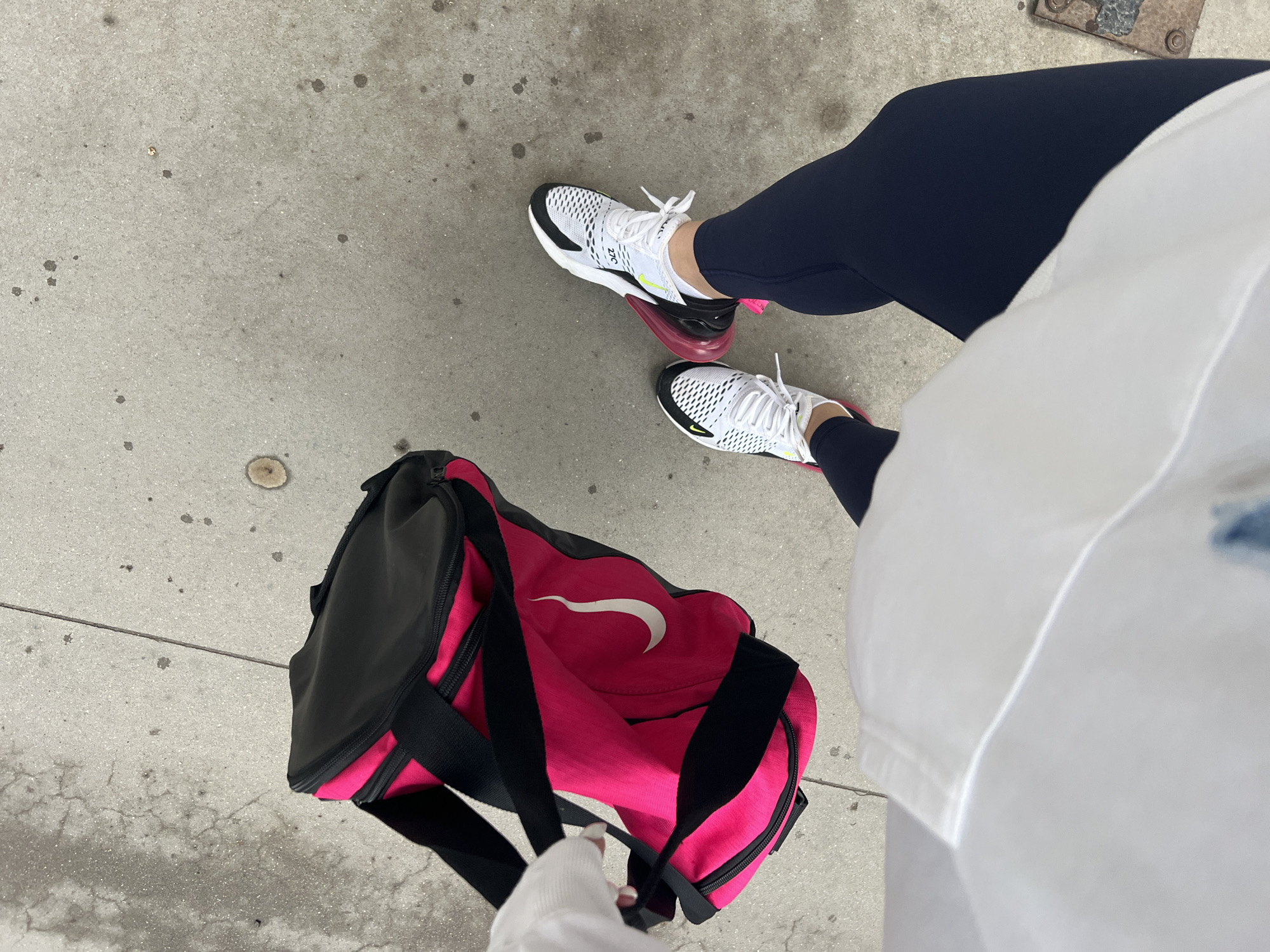 The image provides a high-angle, overhead perspective of a person's lower body and an athletic bag on concrete pavement in Plovdiv, Bulgaria. The person is wearing dark navy blue athletic leggings and white Nike Air Max 270 sneakers, which feature black accents, white laces, and a prominent bright pink air sole unit. The text "270" is visible on the side of the shoes. A white top or jacket is partially visible on the right side of the frame, covering the upper body, and one hand, with light nail polish, holds a strap of the duffel bag.
To the left, a bright pink and black duffel bag rests on the ground. It features black trim, zippers, and straps, with a clear white Nike swoosh logo displayed on its side. The concrete pavement beneath shows signs of wear with small dark stains and minor cracks, indicative of an outdoor urban or suburban setting. A small, partially obscured metal grate or cover is visible in the upper right corner of the frame. The lighting is natural and diffused, suggesting it is daytime, possibly under an overcast sky or in a shaded area. The scene depicts an individual either arriving at or departing from a physical activity, given the athletic attire and sports bag.