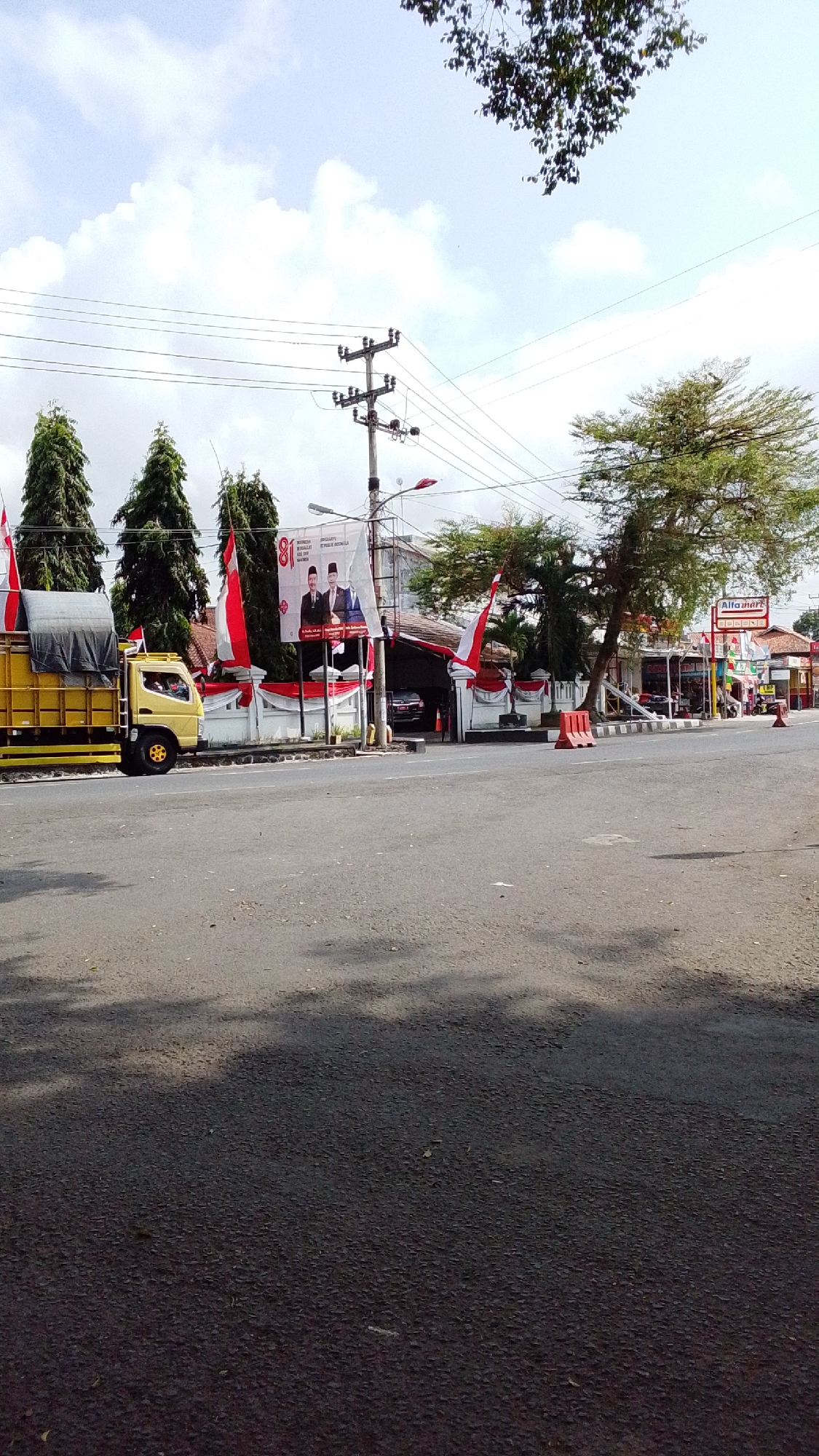 This image captures a street scene in Desa Karangbenda, Indonesia, likely during a celebration, given the prominent red and white flags and banners. On the left, a yellow truck with a gray tarp-covered load is parked on the side of the road. In the background, a building complex is decorated with red and white draped fabric and several large banners. One banner prominently displays the number "81" alongside images of two men in formal attire. Several tall, slender evergreen trees stand in a row, interspersed with more red and white flags. A utility pole with electrical wires runs across the sky. To the right, an Alfamart convenience store is visible, with more flags and decorations. The road surface is asphalt, and shadows from trees are cast across it. The sky is partly cloudy. The scene is a public street with commercial establishments and residential elements.