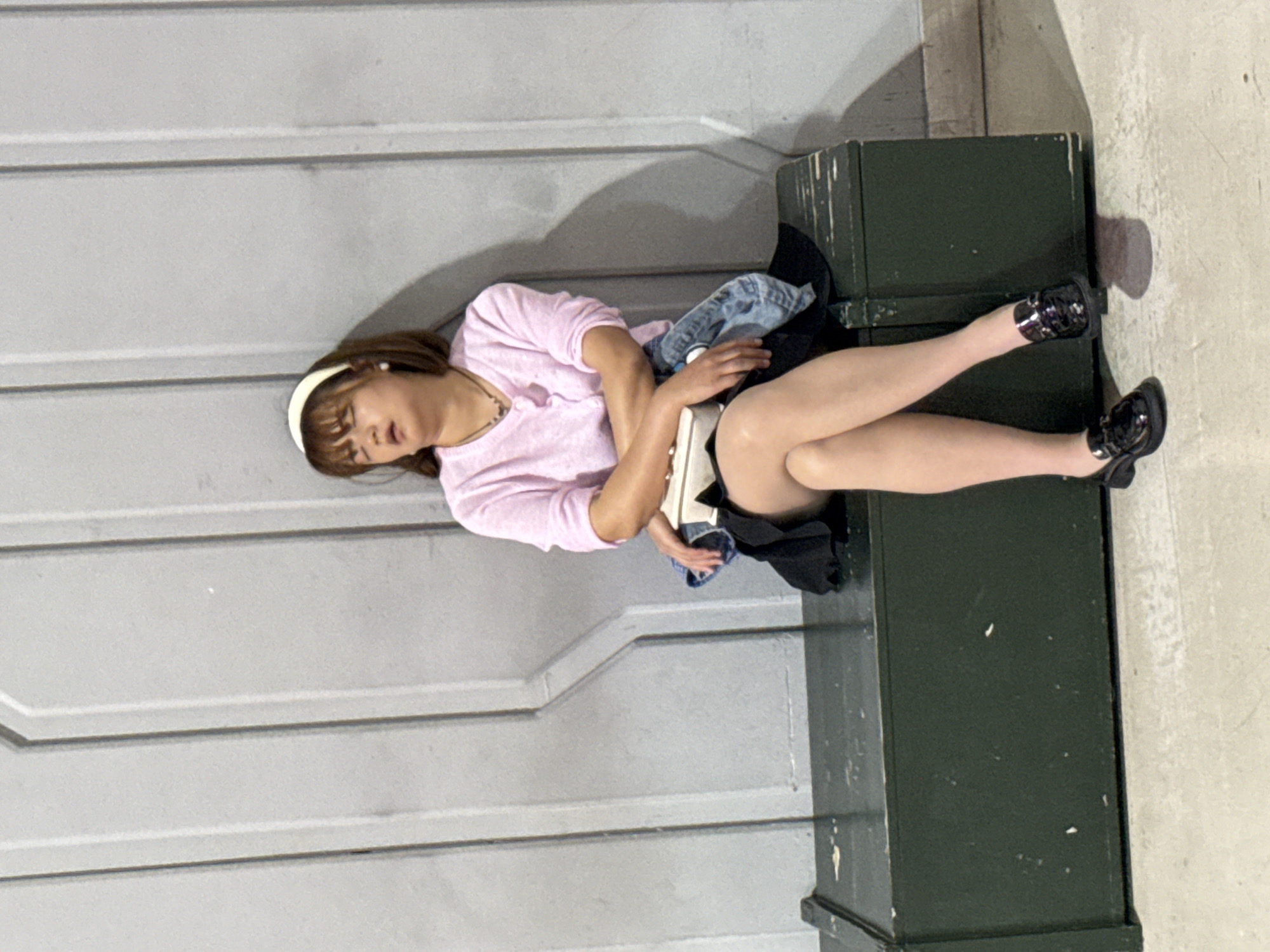 A woman wearing a pink shirt, black skirt, black shoes, and a white headband is lying down. Her head rests on a gray metal surface, and her legs are propped up on a green metal object. She is holding a denim jacket. The location is likely江北城街道, China.