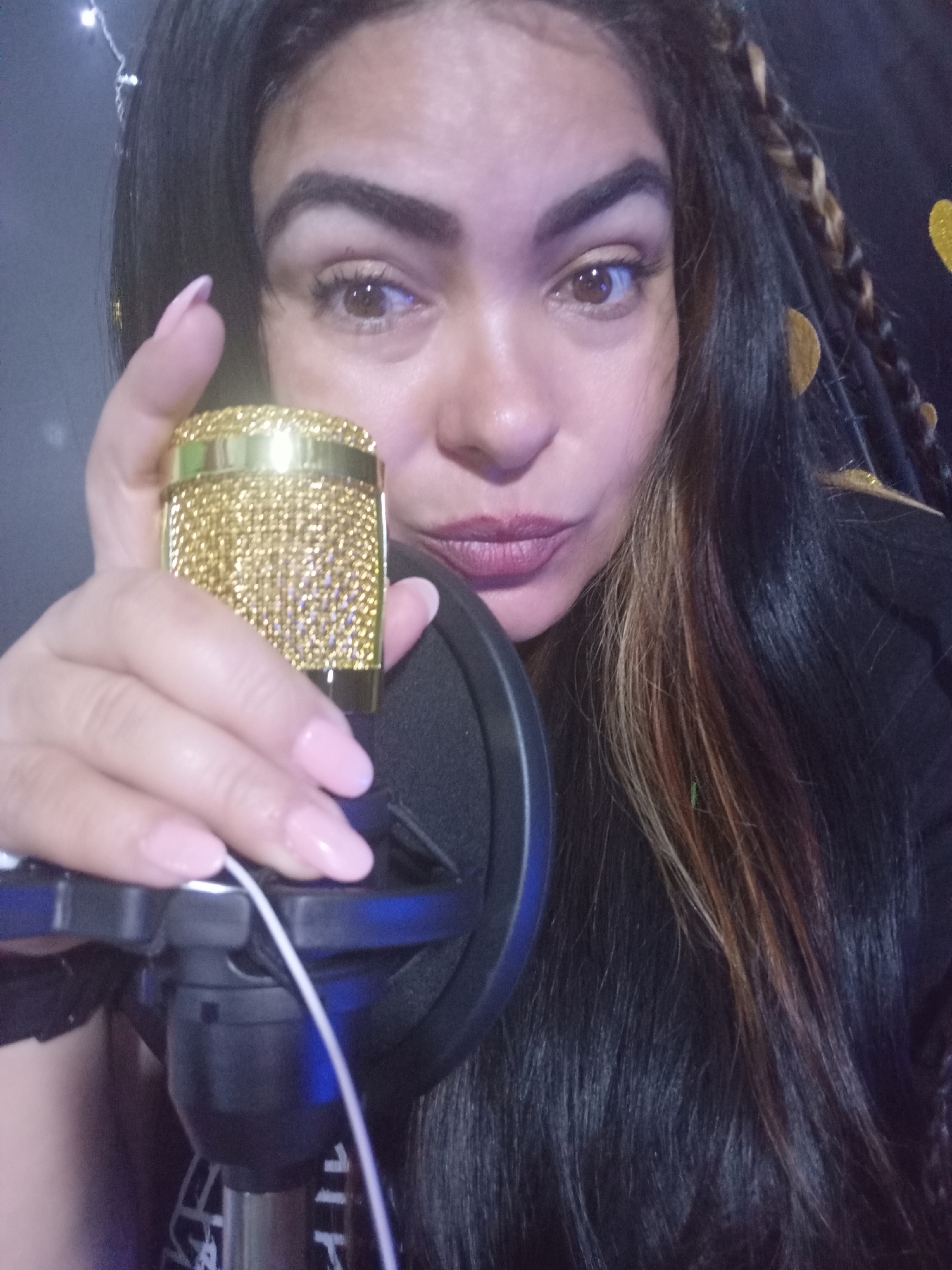 A woman with long dark hair with some blonde streaks and braids is holding a gold microphone with a black pop filter in front of her face. Her nails are painted a light pink. The background is dark with some faint gold decorative elements. There is no indication of this photo being taken in Juan Griego, Venezuela.