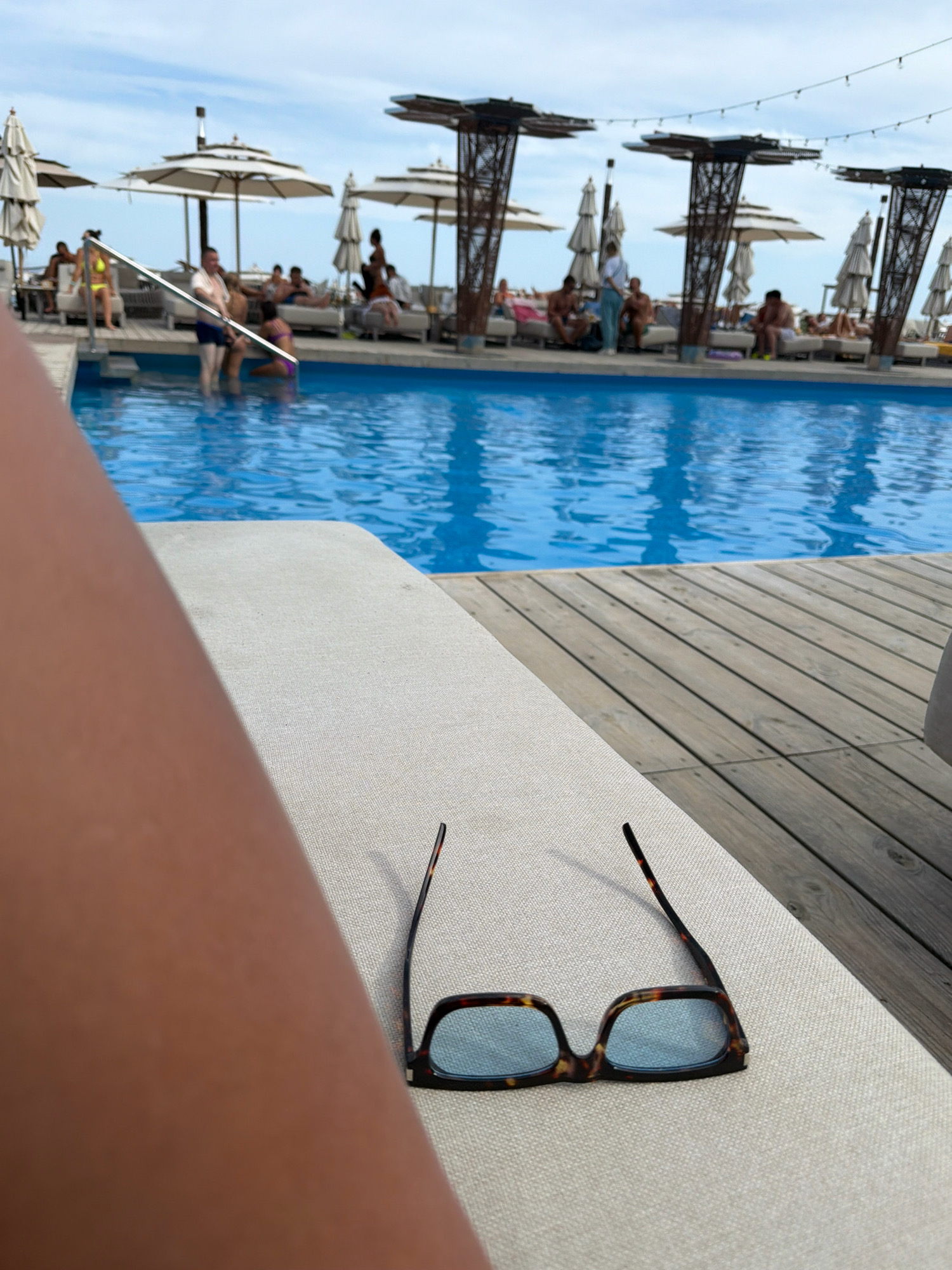An outdoor scene features a swimming pool with clear blue water and a wooden deck. In the foreground, on a light-colored lounge chair, a pair of tortoise-shell framed glasses with light blue lenses rests. The left side of the image is dominated by the blurred silhouette of a person's arm and leg. In the background, several people are visible lounging on chairs and standing near the pool. White umbrellas provide shade for some areas. The sky is partly cloudy, with a bright, hazy atmosphere suggesting it is daytime. The overall environment indicates a recreational resort or beach club setting. The provided information states the location is Odesa, Ukraine.