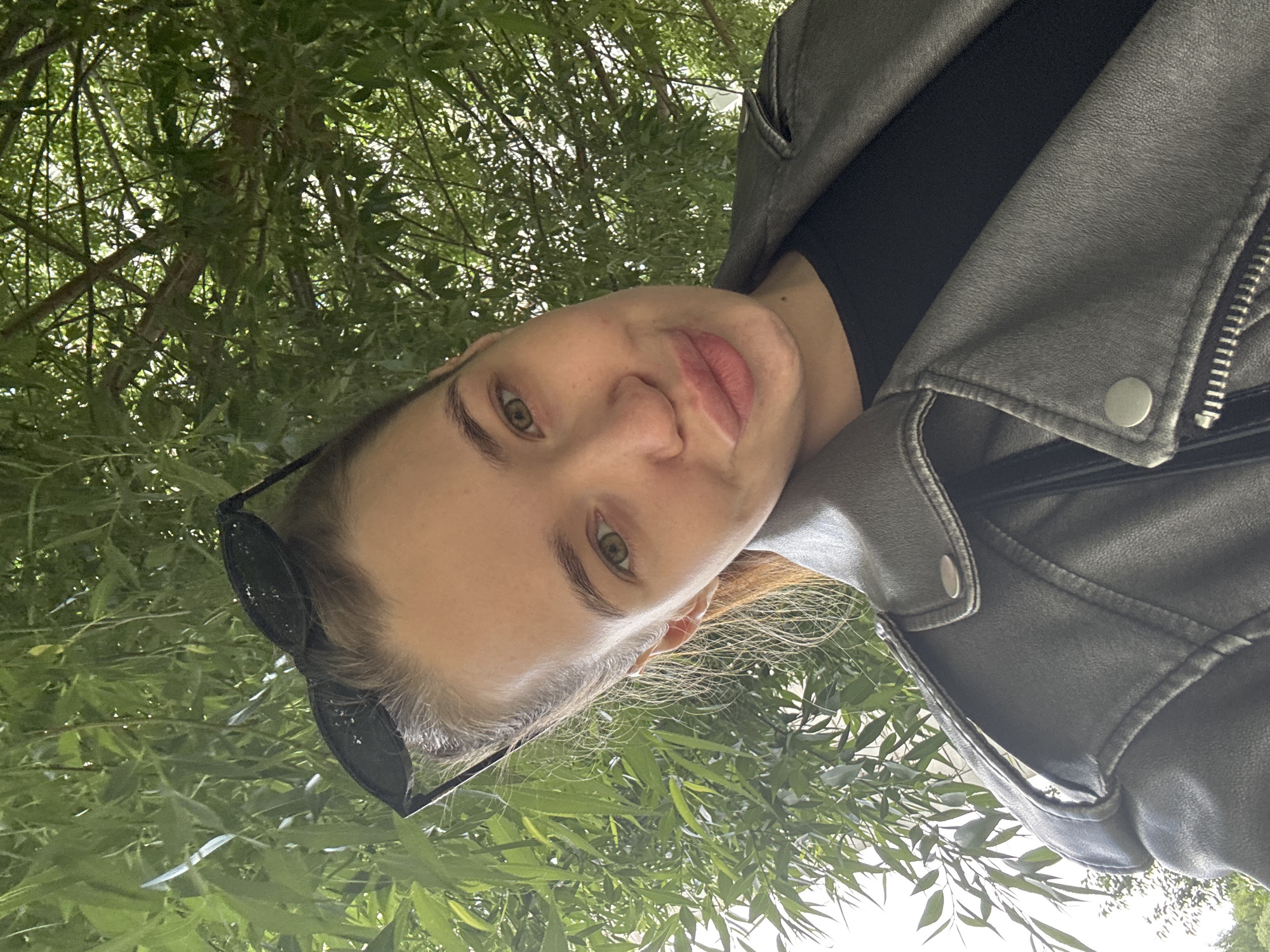 A woman is seen in a close-up selfie, taken from a low angle looking up. She has fair skin, light brown hair, and green eyes. Her lips are full and naturally colored. She is wearing a black top under a gray leather jacket. Sunglasses are perched on top of her head. The background is filled with green foliage, possibly trees, creating a canopy-like effect above her. The location cannot be determined to be the city of Chaikovsky, Russia, from the image alone.
