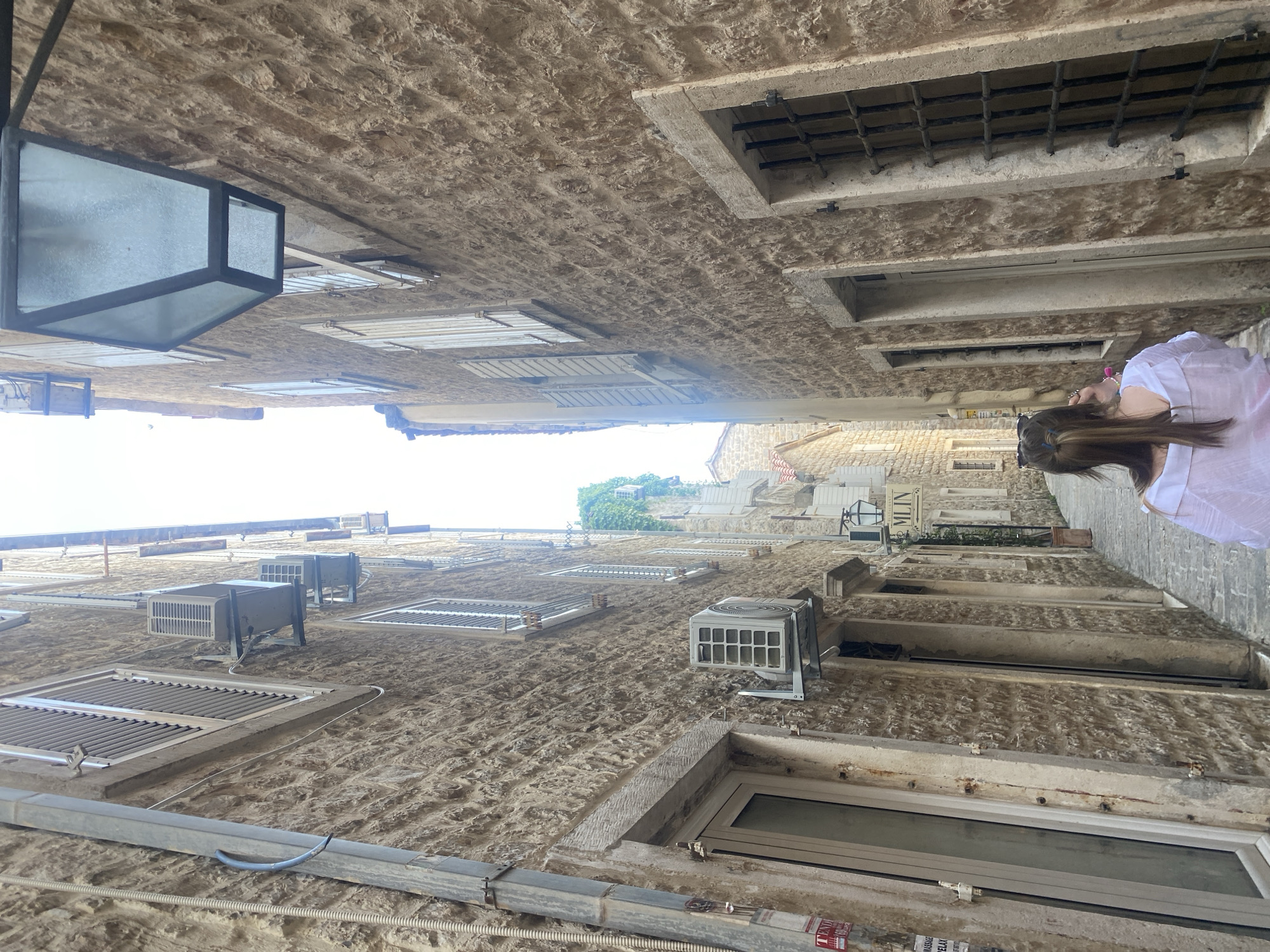 The image depicts an alleyway in Budva, Montenegro, looking up towards the sky. Buildings made of roughly hewn stone line either side. Windows of varying sizes and styles are visible, some with metal grates or slats, and some are modern. Air conditioning units are installed on the walls. A black, boxy streetlamp hangs on the left. On the right, a person with long brown hair and wearing a white shirt is visible, looking upwards toward the buildings. The perspective is from a low angle, looking directly up.