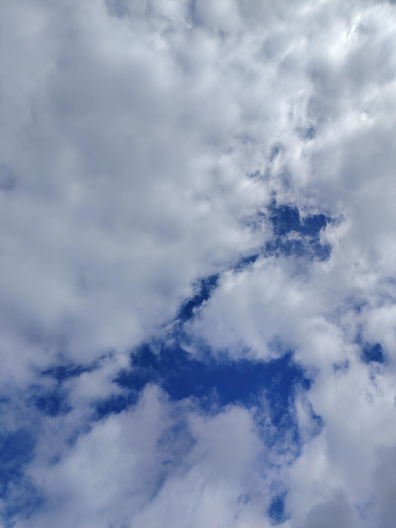 The image is a close-up, aerial shot of a cloudy sky over Хоругвино, Russia. The clouds are predominantly white and light gray, with some areas of darker gray suggesting depth and thickness. Patches of bright blue sky are visible through breaks in the clouds. The lighting suggests it is daytime, with sunlight filtering through the clouds, creating a slightly diffused effect. There are no people, man-made objects, or discernible ground features visible in this image.