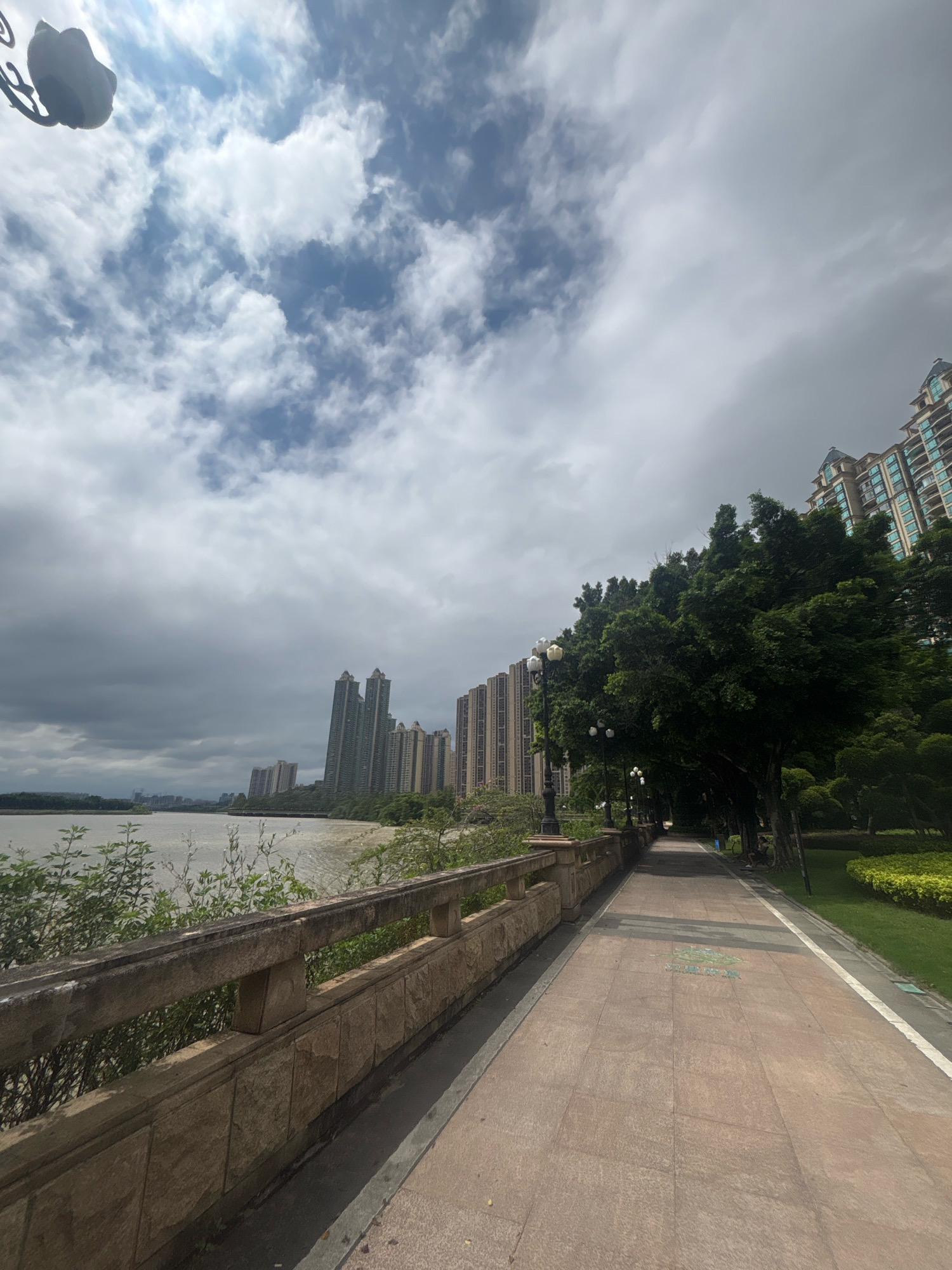 The image depicts a paved walkway alongside a body of water in Nanhai District, China. The walkway is bordered by a stone railing on the left and lush green trees and manicured bushes on the right. Tall apartment buildings are visible across the water, and a line of trees also runs along the water's edge. The sky is partly cloudy, with a mix of blue sky and gray clouds. The overall atmosphere appears to be calm and serene, suggesting a public park or waterfront area designed for recreation. There are no people visible in the image.